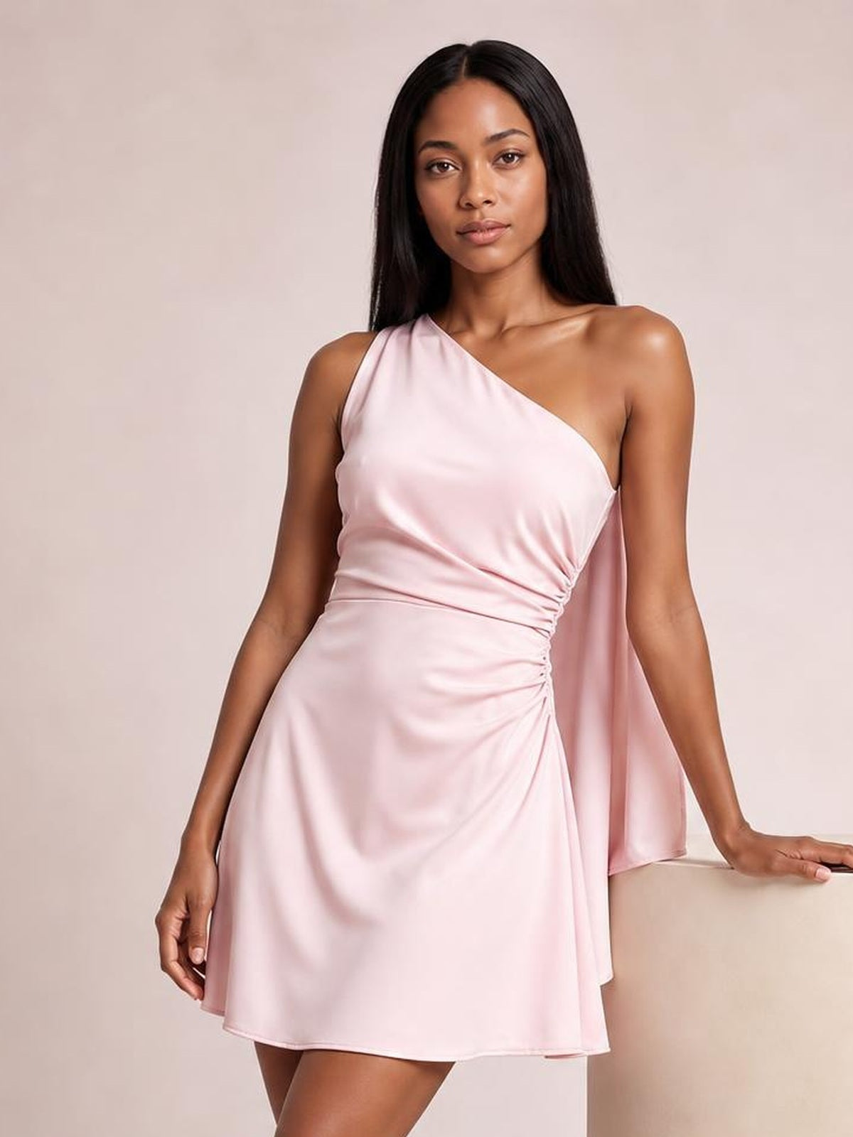 A model poses in a pale pink satin one-shoulder mini dress with a draped side panel, ruched waist, asymmetrical neckline, and fluid A-line silhouette.