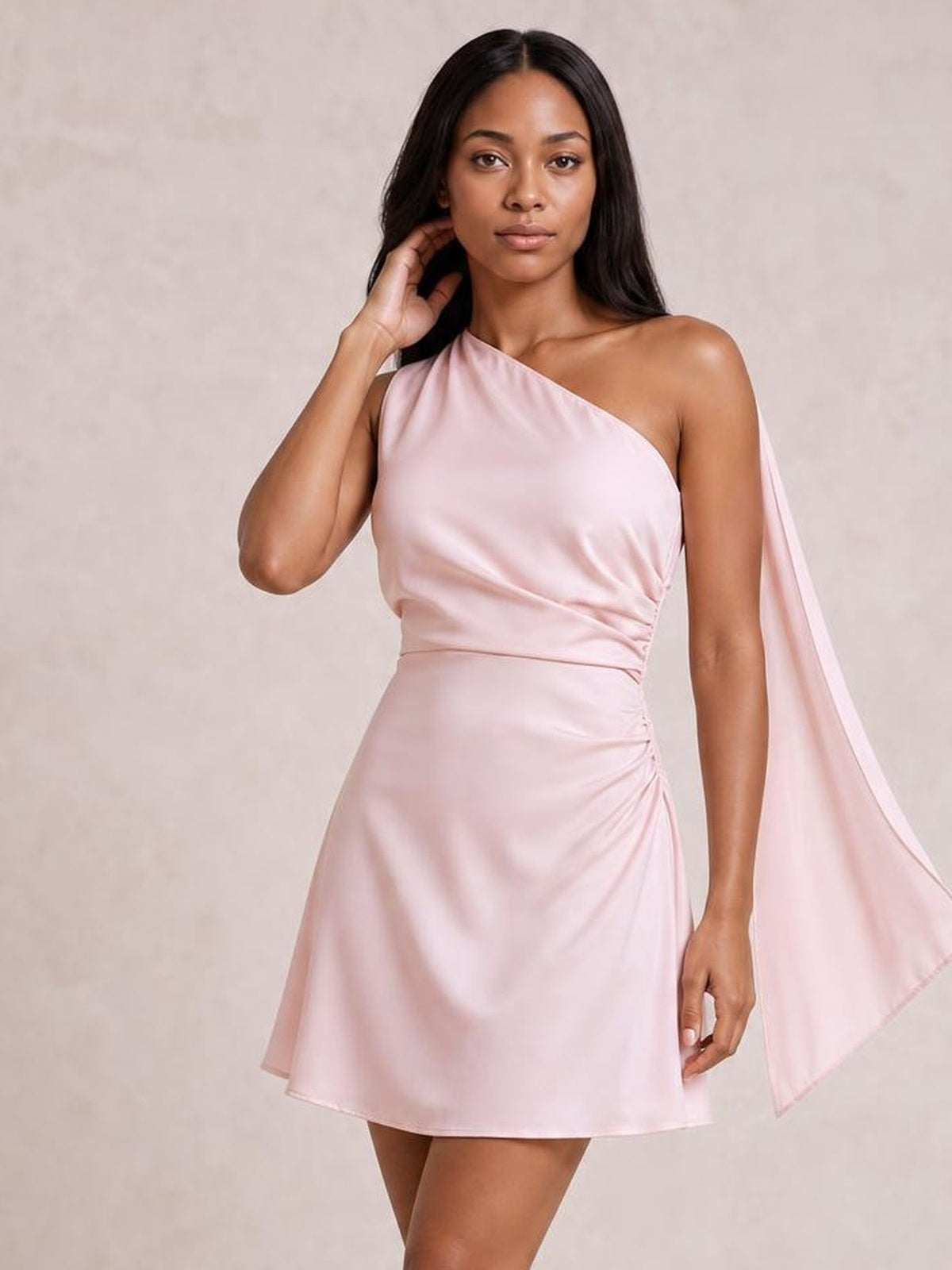 Model poses in a pale pink satin one-shoulder mini dress with a draped shoulder detail, ruched waist, fitted silhouette, and flared hem.