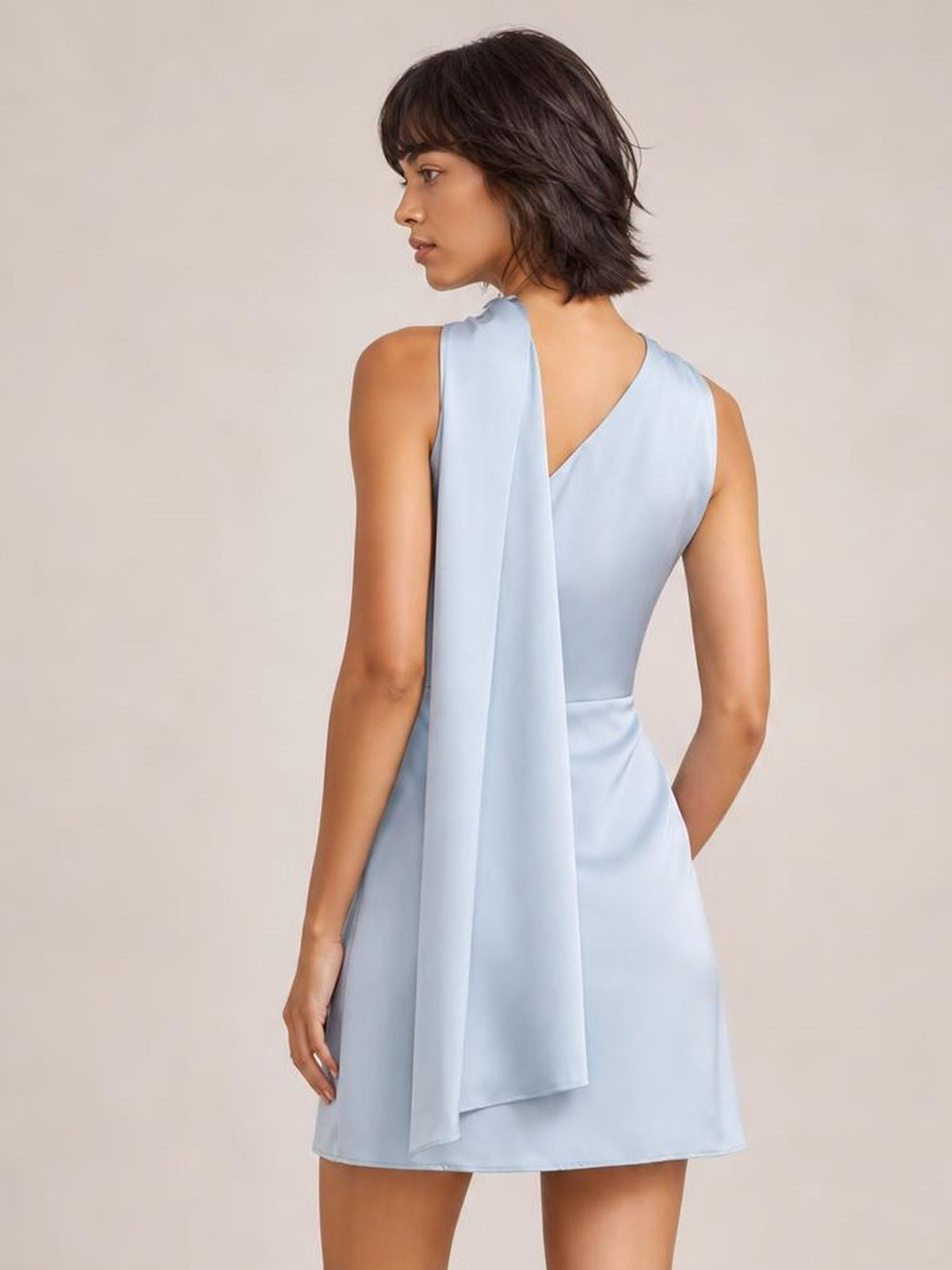 Model turns to show the back of a light blue satin one-shoulder mini dress with a long draped panel, ruched shoulder detail, and softly flared silhouette.