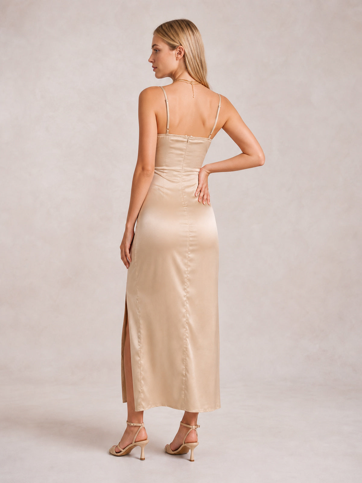 A model poses with one hand on her hip, showing a champagne satin spaghetti strap midi dress with a fitted silhouette, back seam, and side slit.