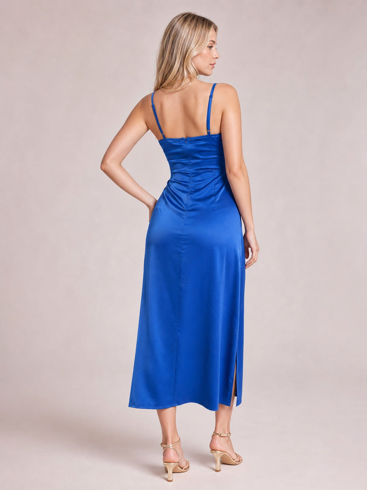 A model poses in a vibrant blue satin spaghetti strap slit midi dress, featuring a fitted ruched bodice, smooth flowing skirt, and back seam detailing.