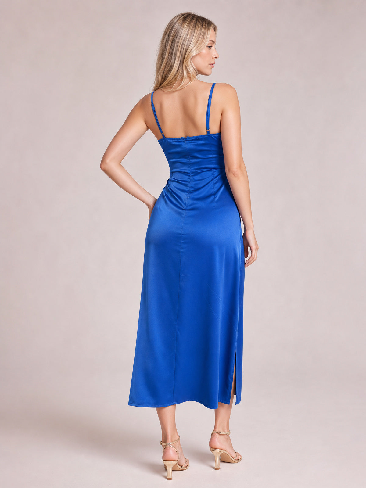 A model poses in a vibrant blue satin spaghetti strap slit midi dress, featuring a fitted ruched bodice, smooth flowing skirt, and back seam detailing.