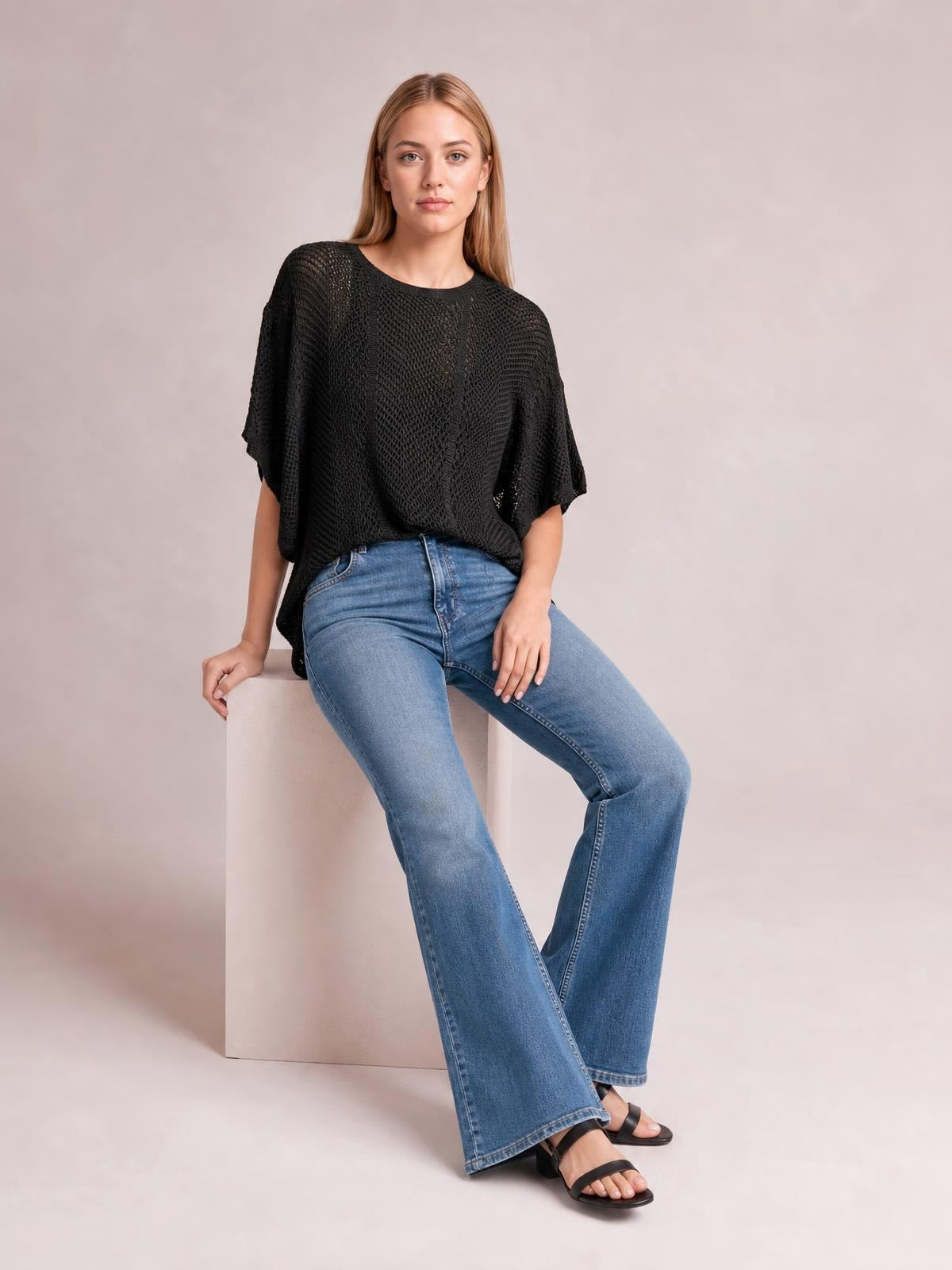 A seated model wears a black scallop-edge half-sleeve pointelle knit top with an openwork texture and relaxed, draped silhouette.