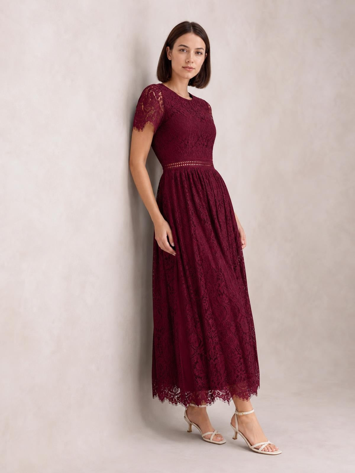 Model poses in a burgundy lace maxi dress with a fitted bodice, defined waist, short lace sleeves, flowing skirt, and scalloped hem trim.