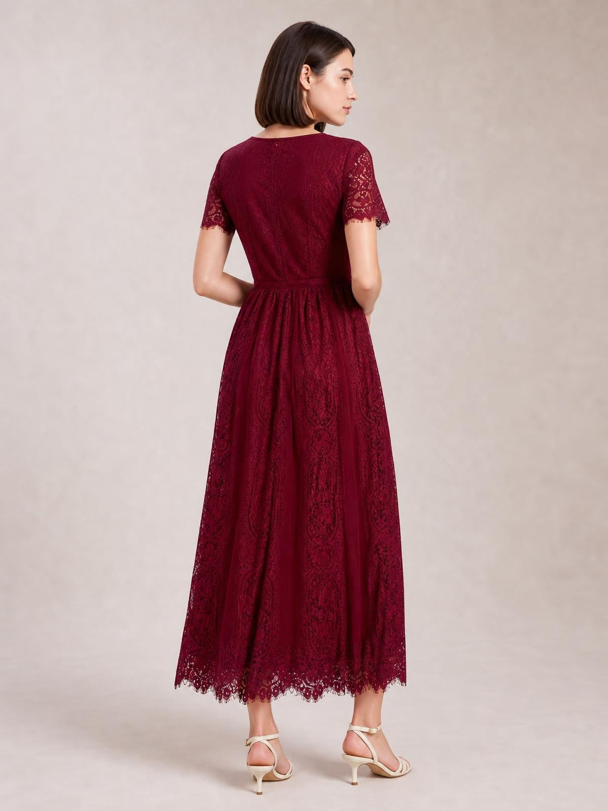 Model stands turned away in a burgundy lace maxi dress with a fitted waist, short scalloped sleeves, flowing A-line skirt, and scalloped hem.