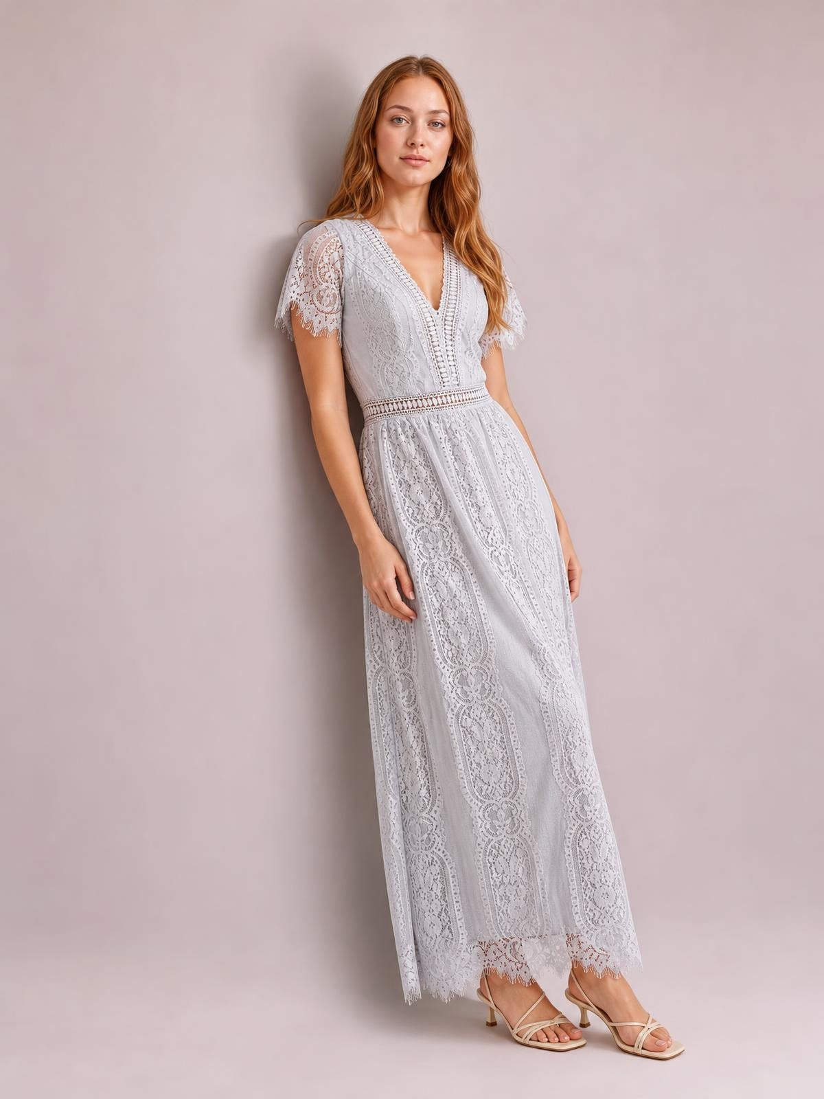 A model leans gracefully in a light gray lace plunge maxi dress with a V-neck, short sleeves, fitted waist, flowing silhouette, and scalloped trim.