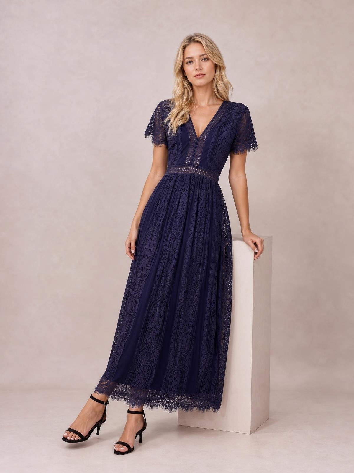 Model stands in a navy lace maxi dress with a plunge V-neck, short scalloped sleeves, defined waistband, and flowing pleated skirt.