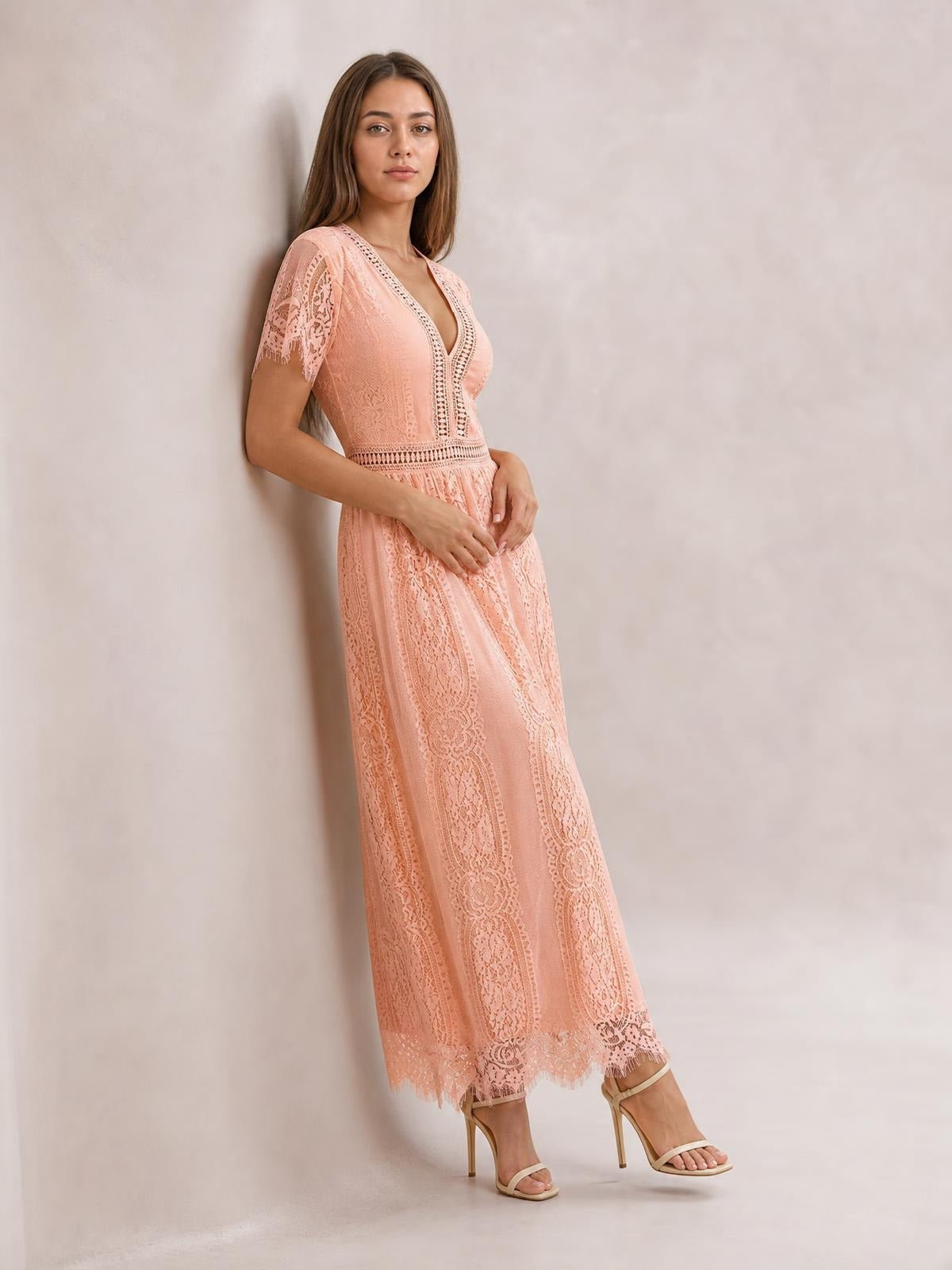 Model leans against a wall in a peach lace maxi dress with a plunging V-neck, short sleeves, defined waist, flowing silhouette, and scalloped hem.