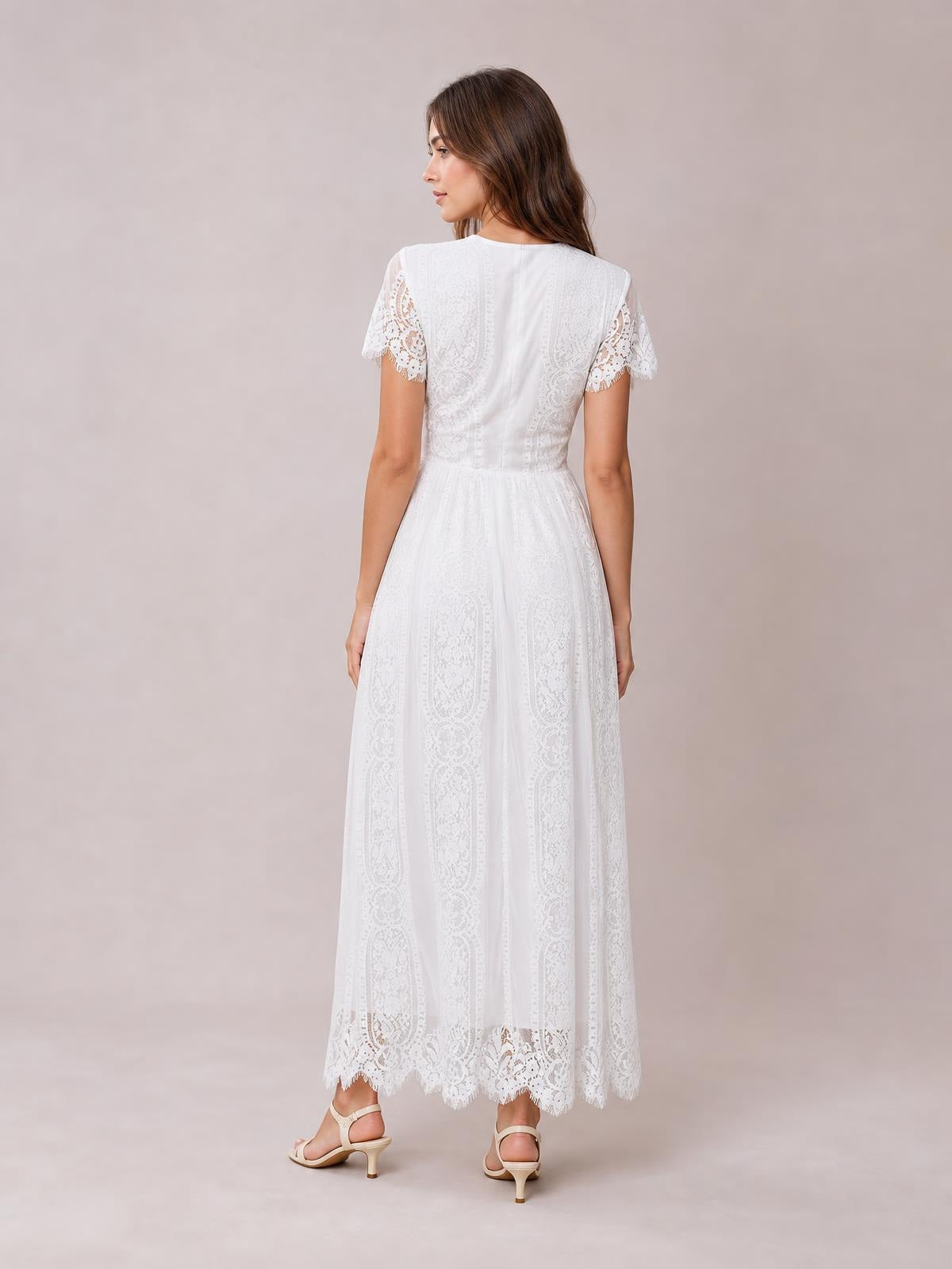 A model faces away wearing a white lace maxi dress with short scalloped sleeves, a fitted bodice, defined waist, and flowing flared skirt with scalloped hem.