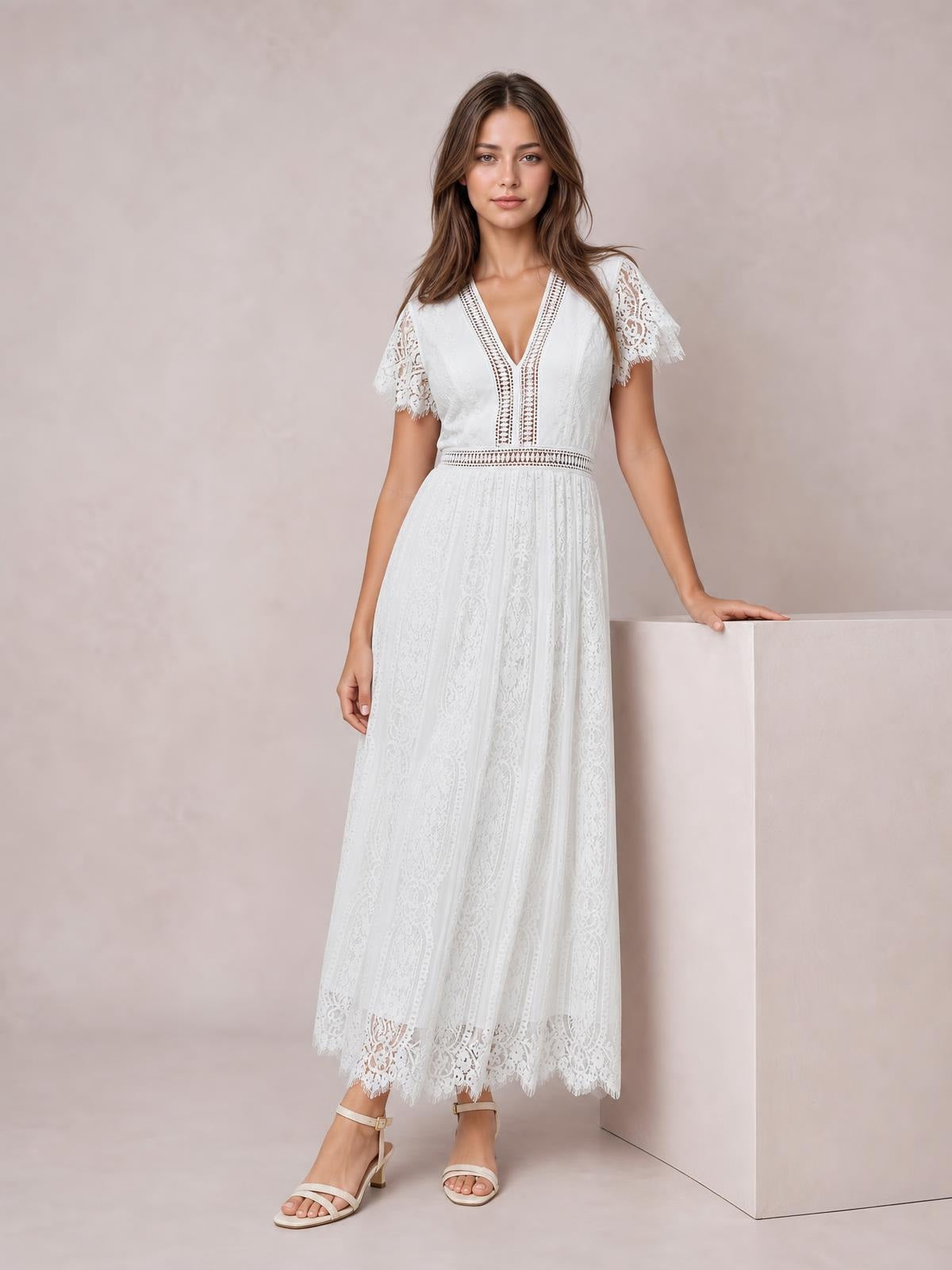 Model poses in a white lace maxi dress with a plunge V-neck, short scalloped sleeves, fitted waist, and flowing A-line silhouette.