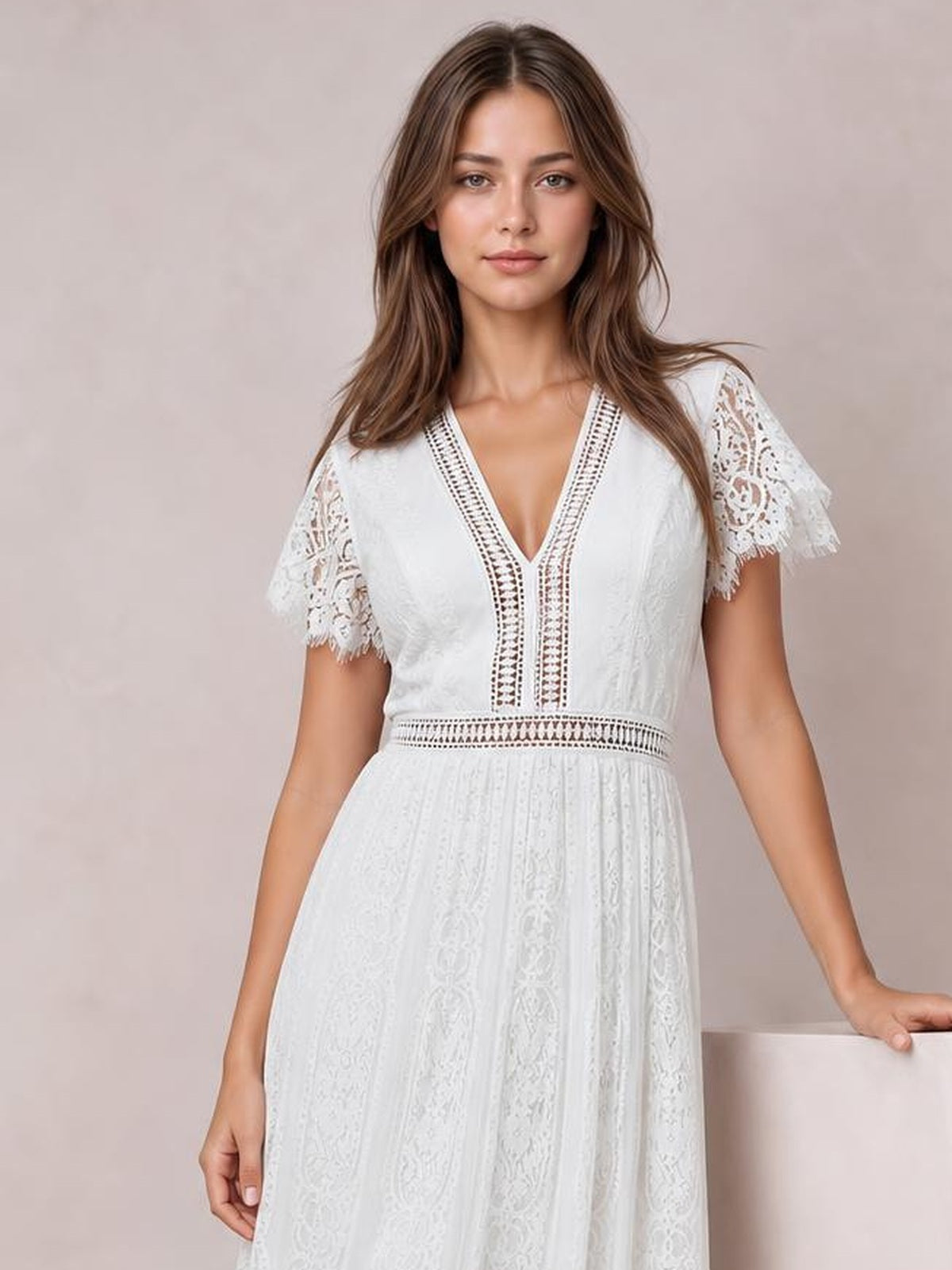 A model stands in a white lace plunge maxi dress with scalloped short sleeves, decorative trim, a defined waist, and a flowing silhouette.