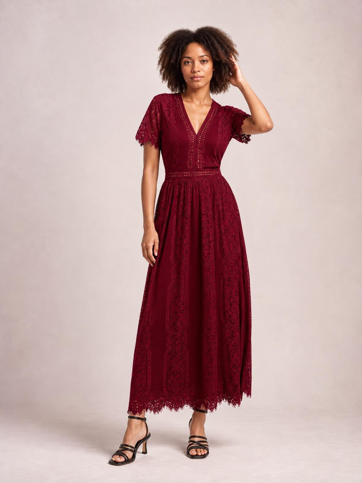 Model poses in a burgundy lace maxi dress with a plunging V-neck, short scalloped sleeves, defined waist, and flowing full-length silhouette.