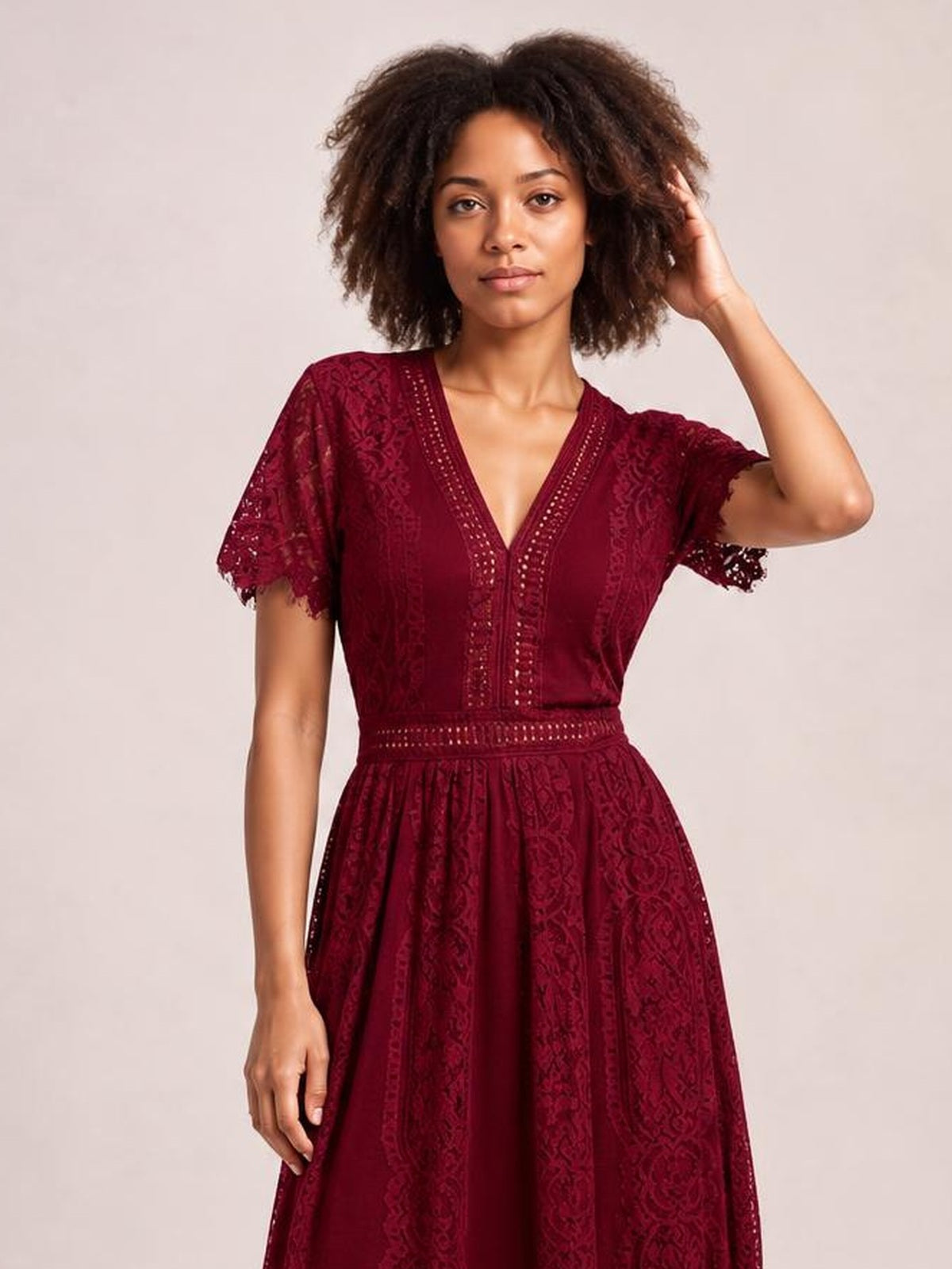 Model wearing a burgundy lace plunge maxi dress with a V-neck, short scalloped-trim sleeves, defined waist, and flowing full skirt, posing with one hand in her hair.