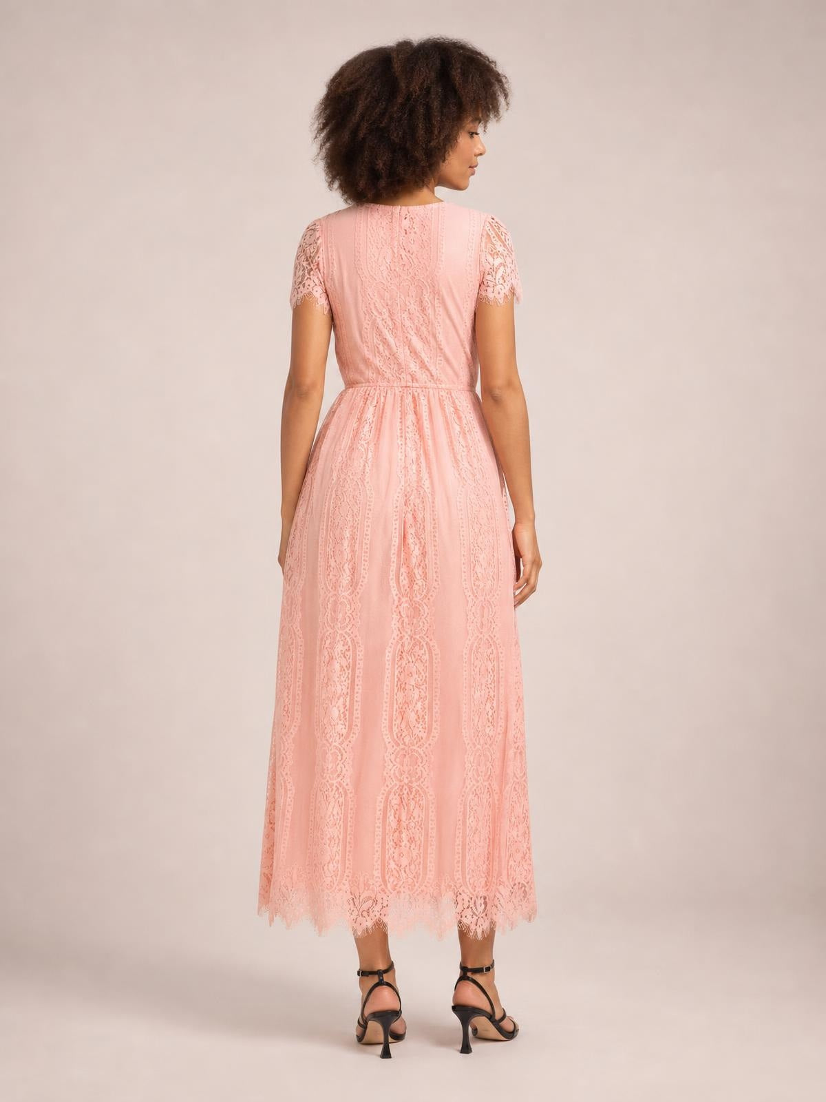 Model stands turned away in a blush pink lace maxi dress with scalloped short sleeves and hem, a fitted waist, and a flowing A-line silhouette.