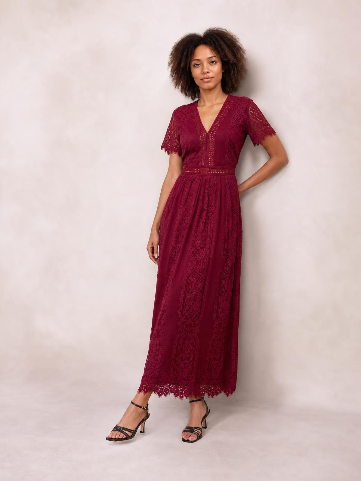 Model poses in a burgundy lace plunge maxi dress with a fitted waist, short scalloped sleeves, flowing silhouette, and scalloped hem.