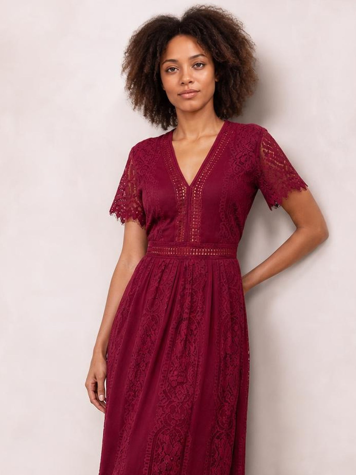 Model poses in a burgundy scalloped trim lace plunge dress with a V-neck, short sleeves, fitted waistband, and flowing maxi silhouette.