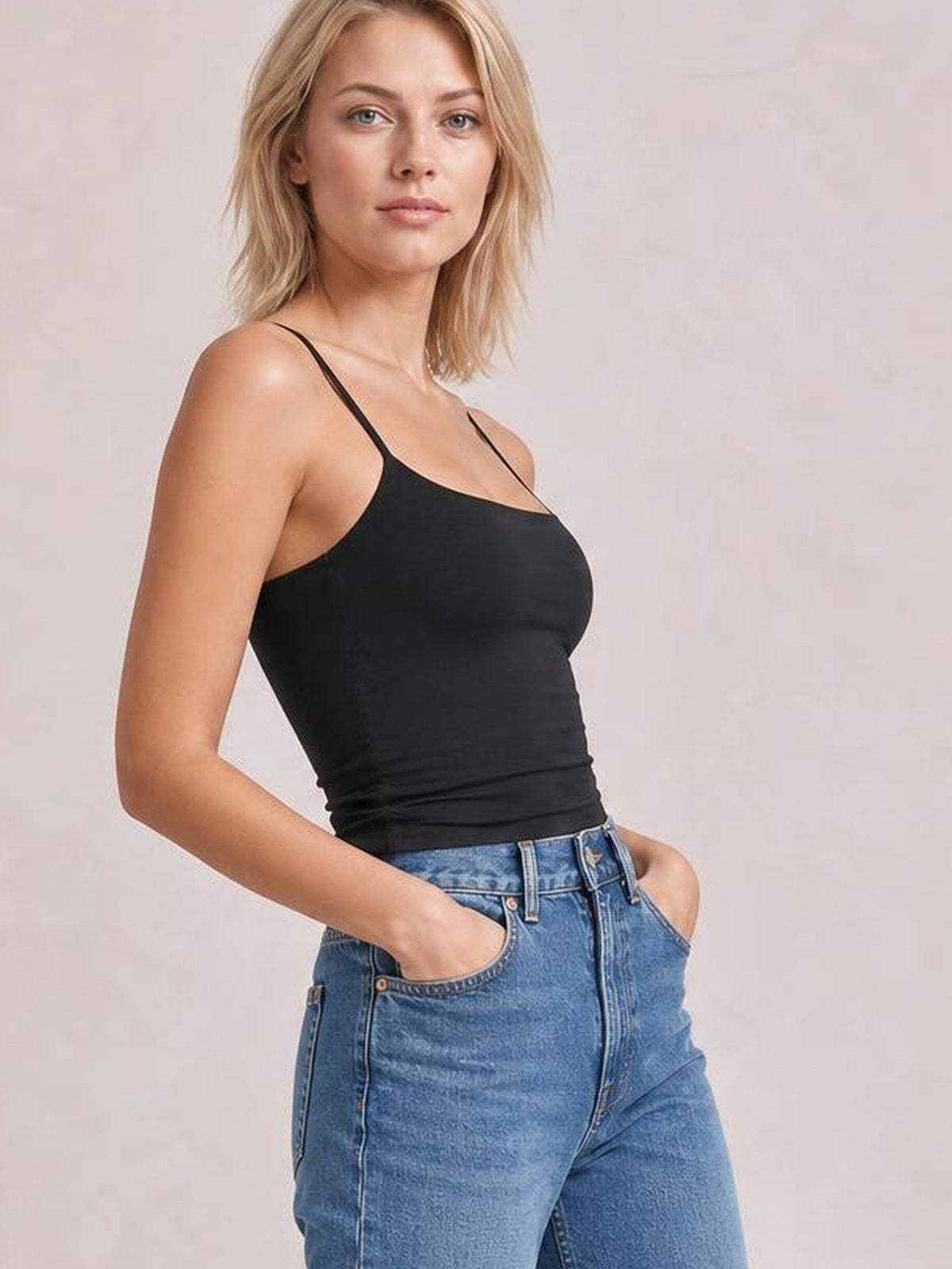 Model wears a black scoop neck cami with thin straps and a fitted, slightly stretchy silhouette, posing with both hands in her jeans pockets.