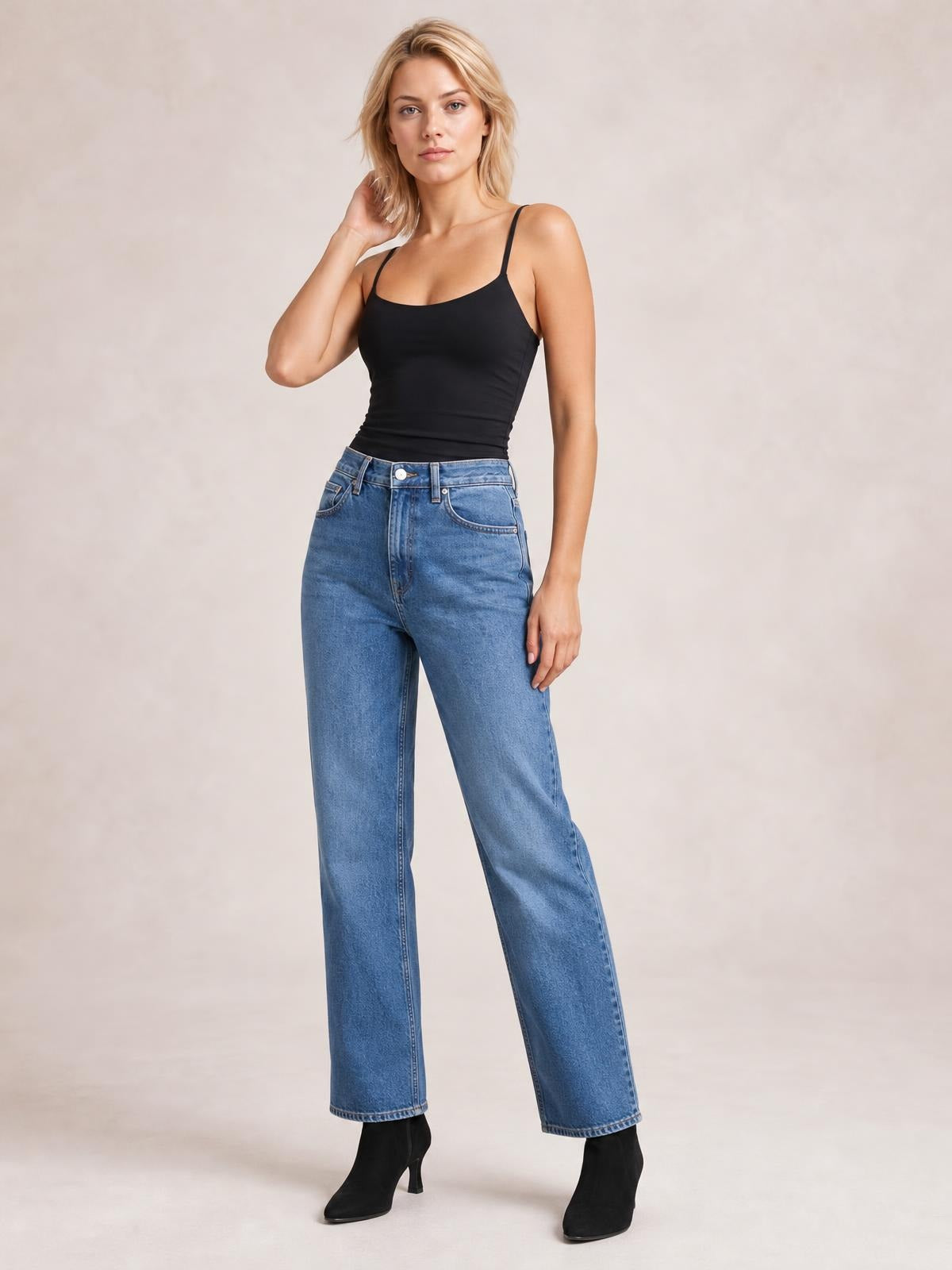 Model poses in a black scoop neck cami with slim straps and a fitted silhouette, styled with high-waisted blue jeans.