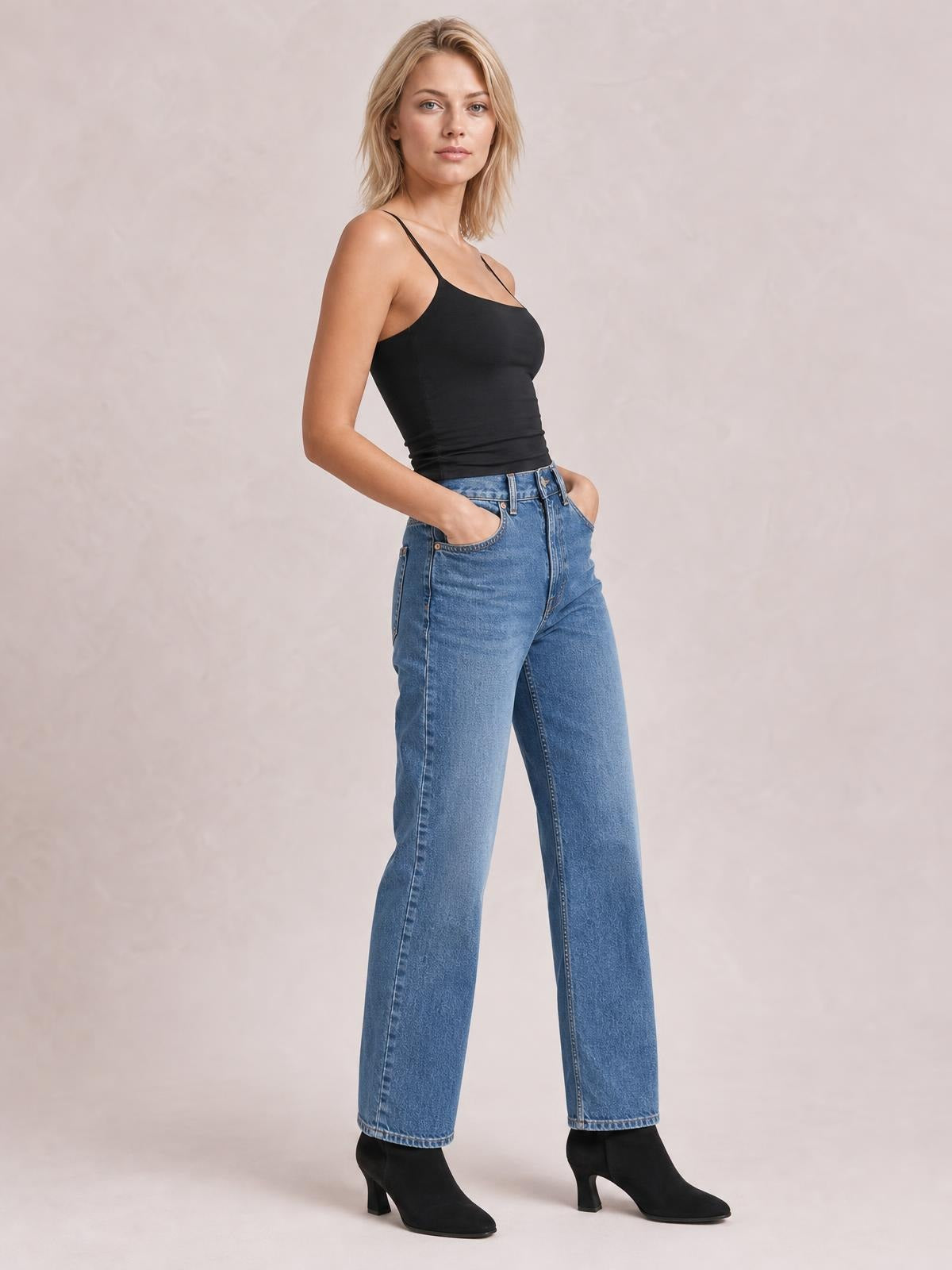 A model poses with hands in her pockets wearing a black scoop neck cami with slim straps and a fitted silhouette, paired with blue wide-leg jeans.