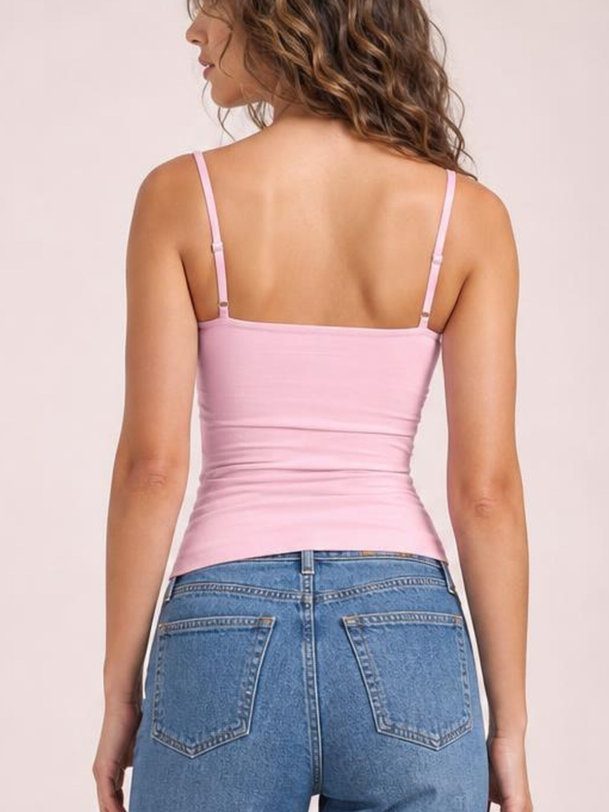 Back view of a model wearing a pink scoop neck cami with thin adjustable straps, a fitted silhouette, and subtle horizontal ruching.