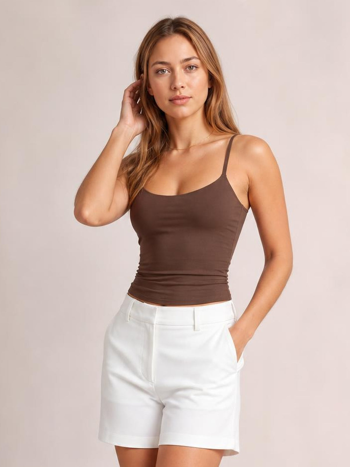 A model poses in a brown scoop neck cami with slim shoulder straps and a fitted, slightly stretchy silhouette, paired with white shorts.