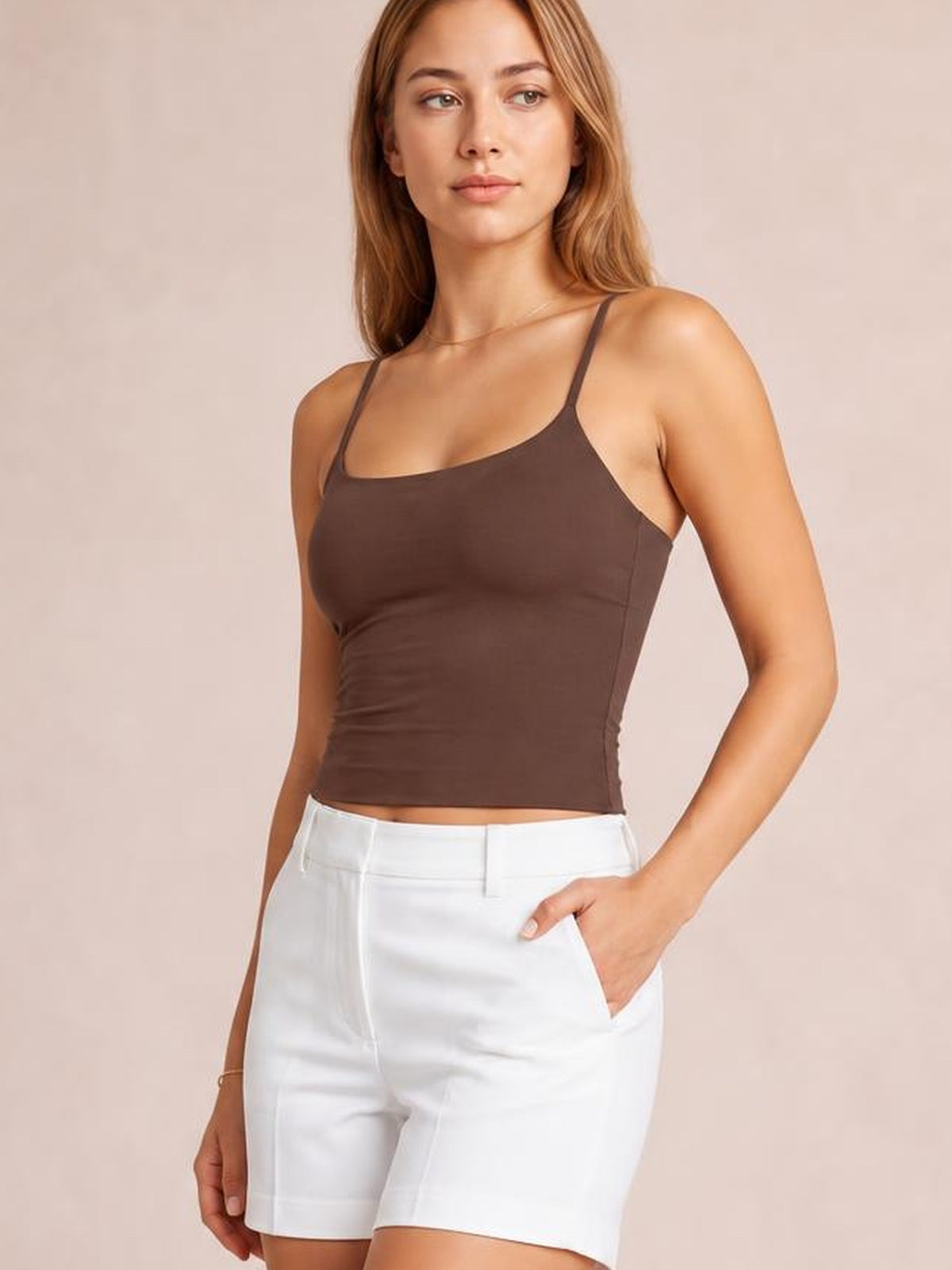 A model wears a chocolate brown scoop neck cami with thin straps and a fitted cropped silhouette, posing with one hand in white shorts.