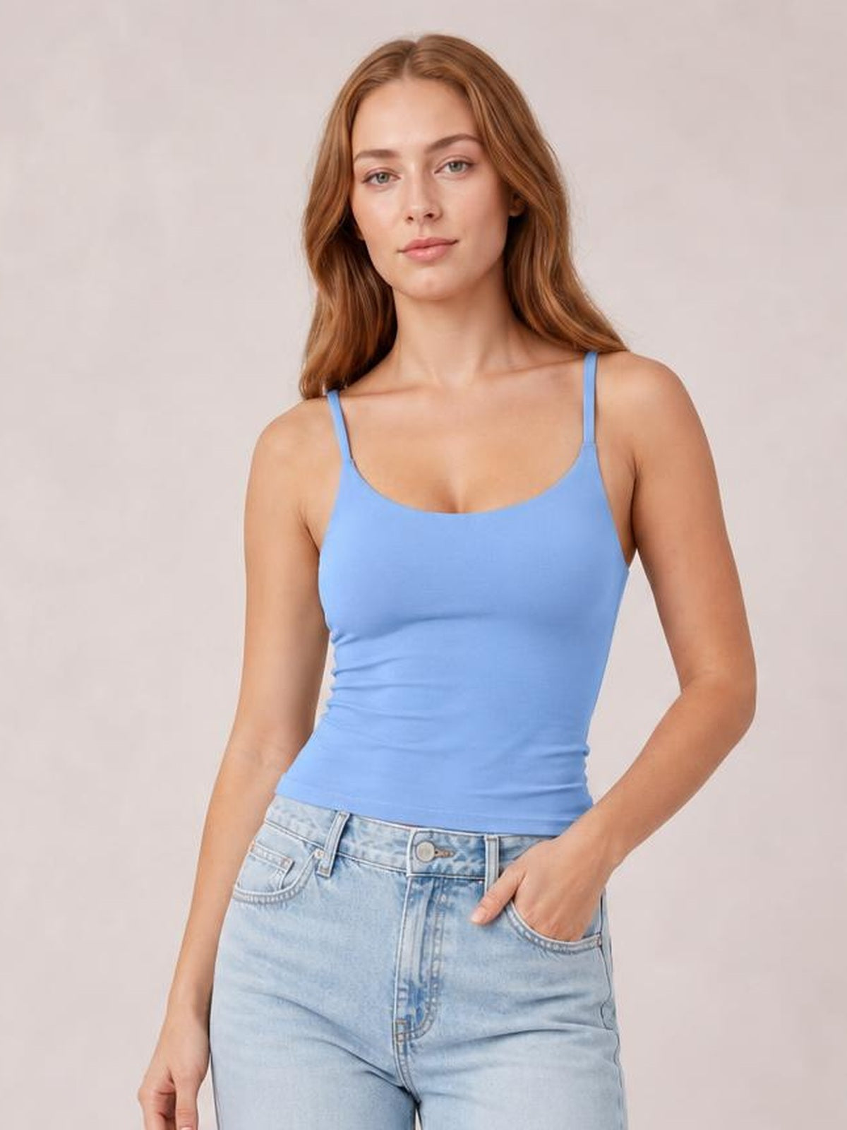 Model wearing a blue scoop neck cami with slim shoulder straps and a fitted, slightly stretchy silhouette, posing with one hand in her jeans pocket.