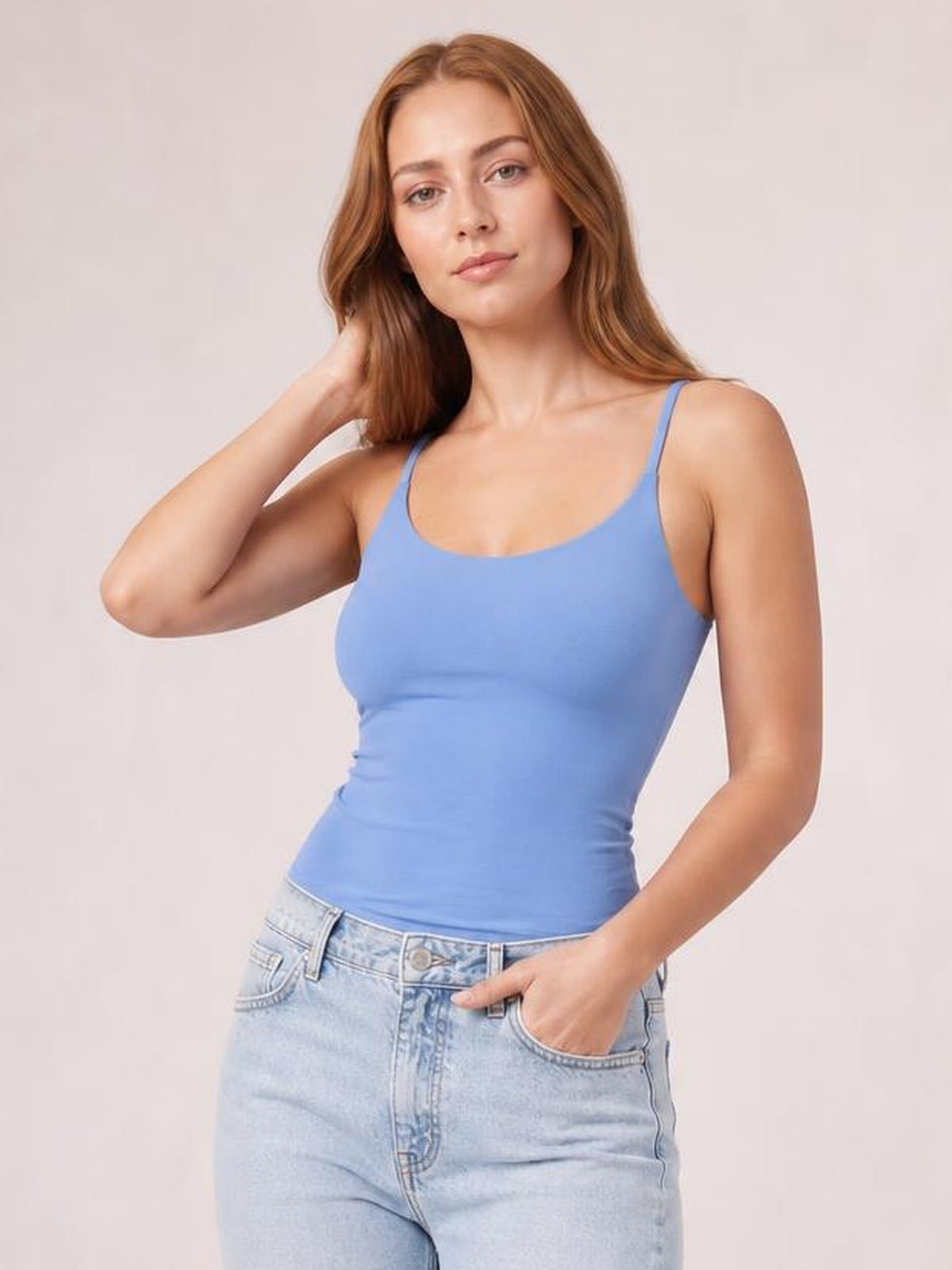 Model poses in a fitted blue scoop neck cami with slim shoulder straps and slight stretch, paired with light-wash jeans.