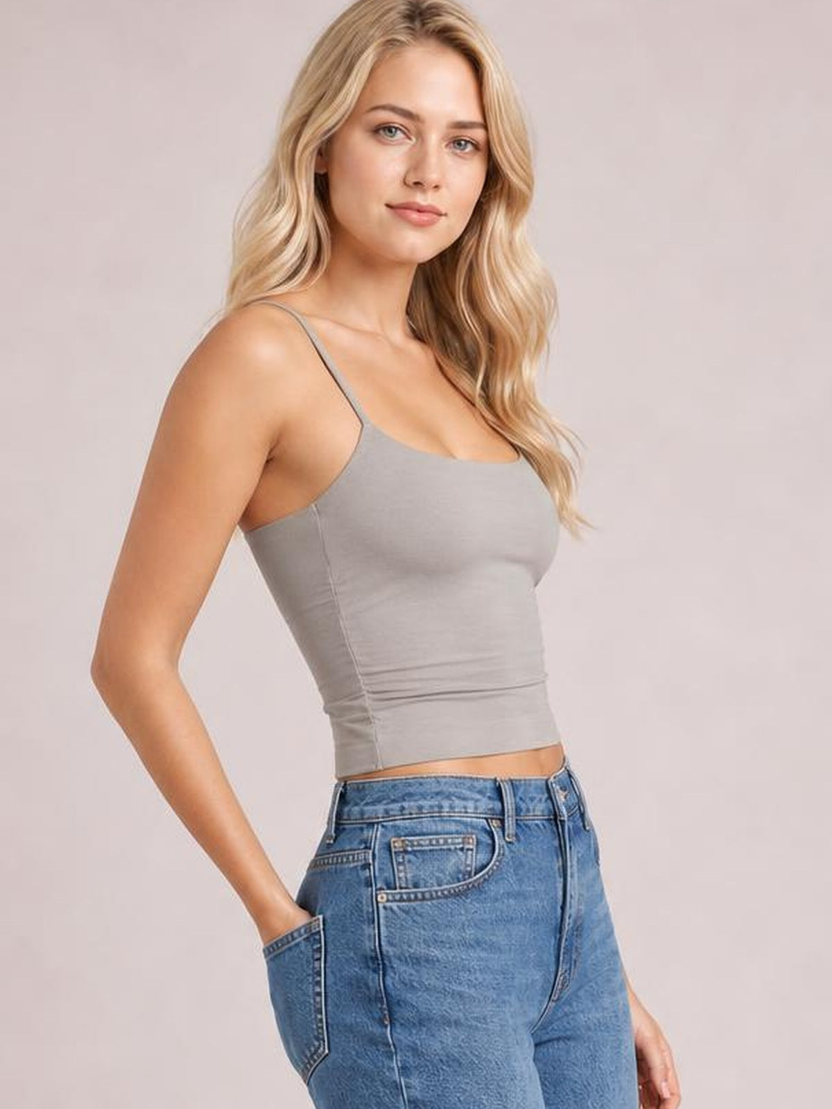 Model poses with one hand in her jeans pocket wearing a fitted gray scoop neck cami with thin straps, a cropped silhouette, and visible side seams.