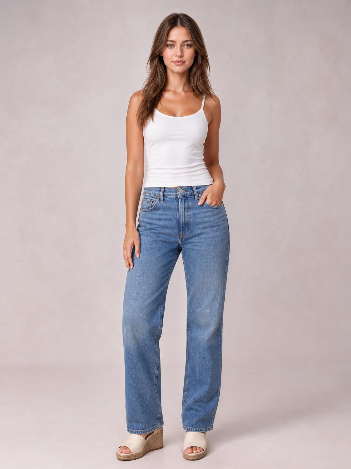 Model poses with one hand in her jeans pocket wearing a white scoop neck cami with thin straps, a fitted silhouette, and subtle stretch.