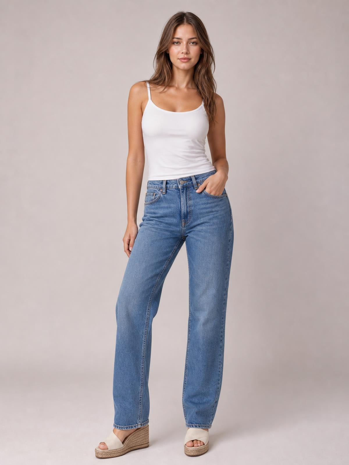 A model stands in a white scoop neck cami with slim straps and a fitted silhouette, paired with relaxed blue jeans, one hand in a pocket.
