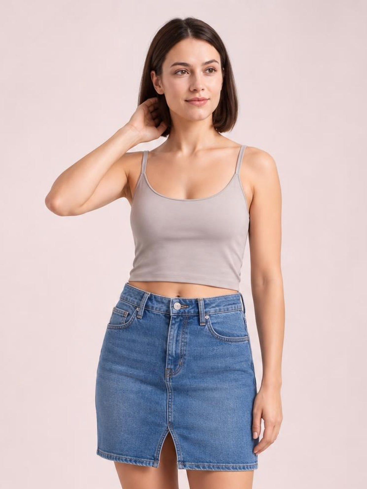 A model poses in a taupe scoop neck cropped cami with thin shoulder straps, a fitted silhouette, and smooth construction, touching her hair.