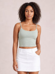 Model wearing a sage green scoop neck cropped cami with thin straps and a fitted silhouette, paired with a white skirt.