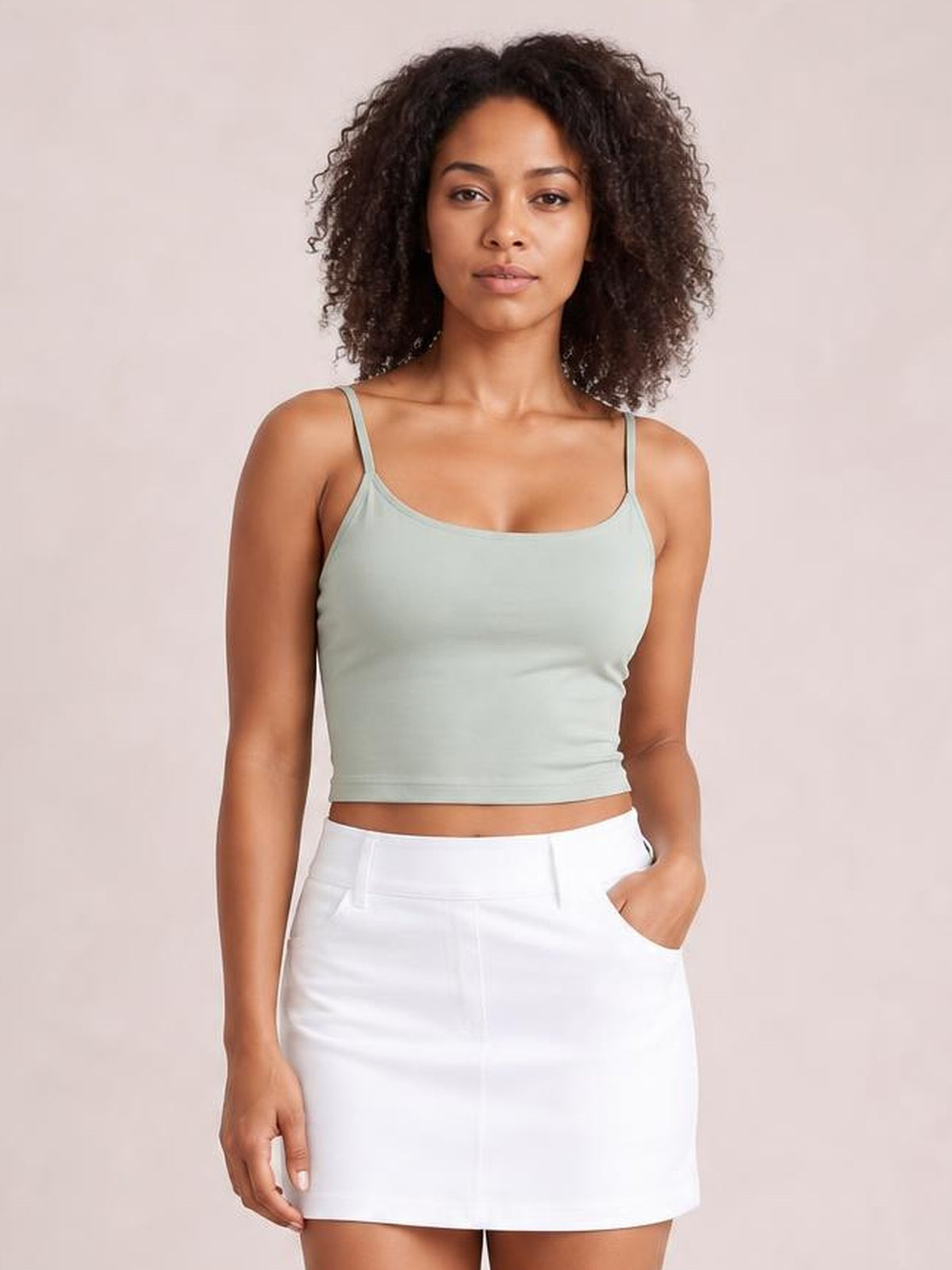Model wearing a sage green scoop neck cropped cami with thin straps and a fitted silhouette, paired with a white skirt.