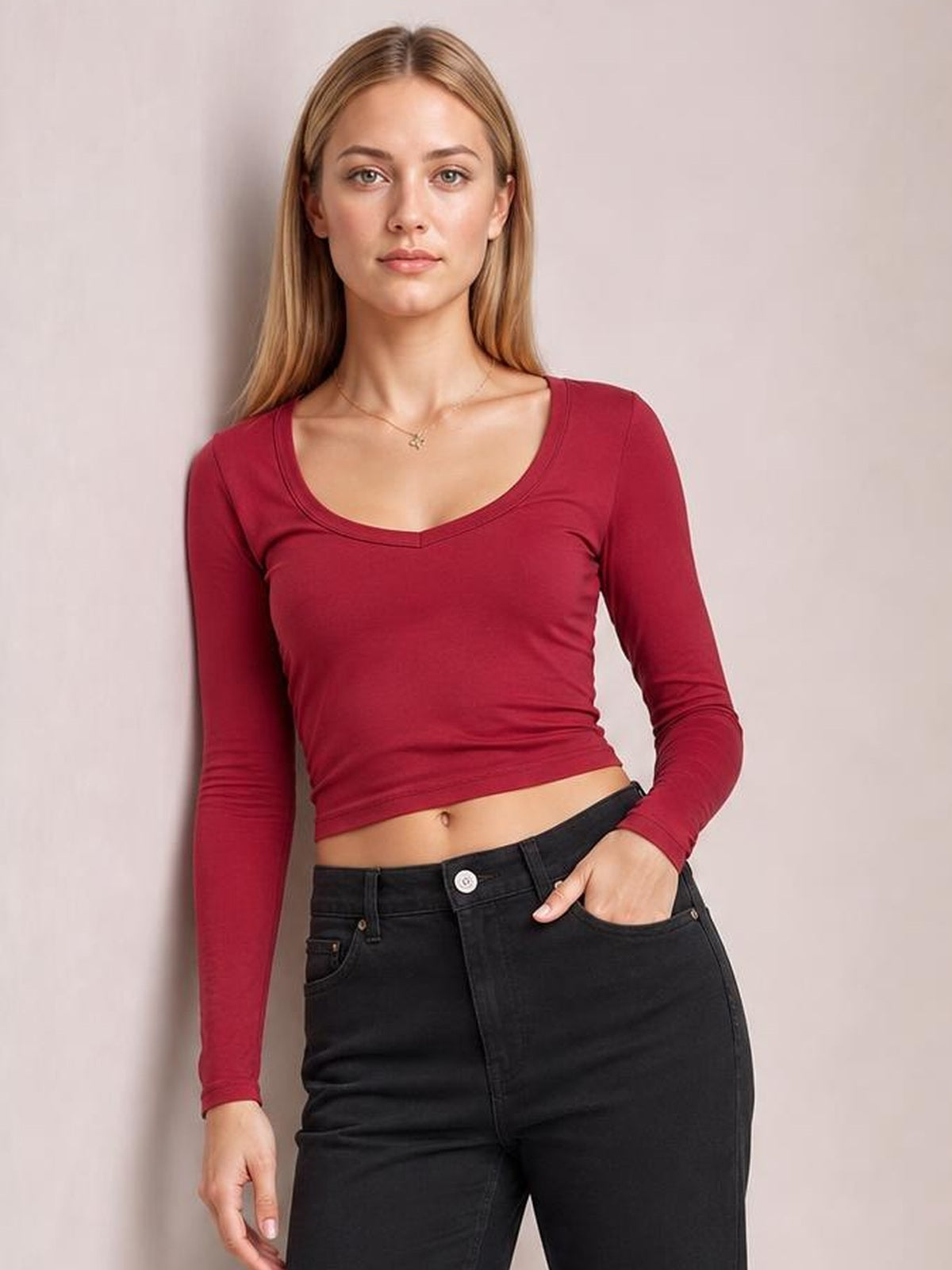 Model poses in a burgundy scoop neck long sleeve cropped T-shirt with a fitted silhouette and visible stitched neckline, one hand in her jeans pocket.