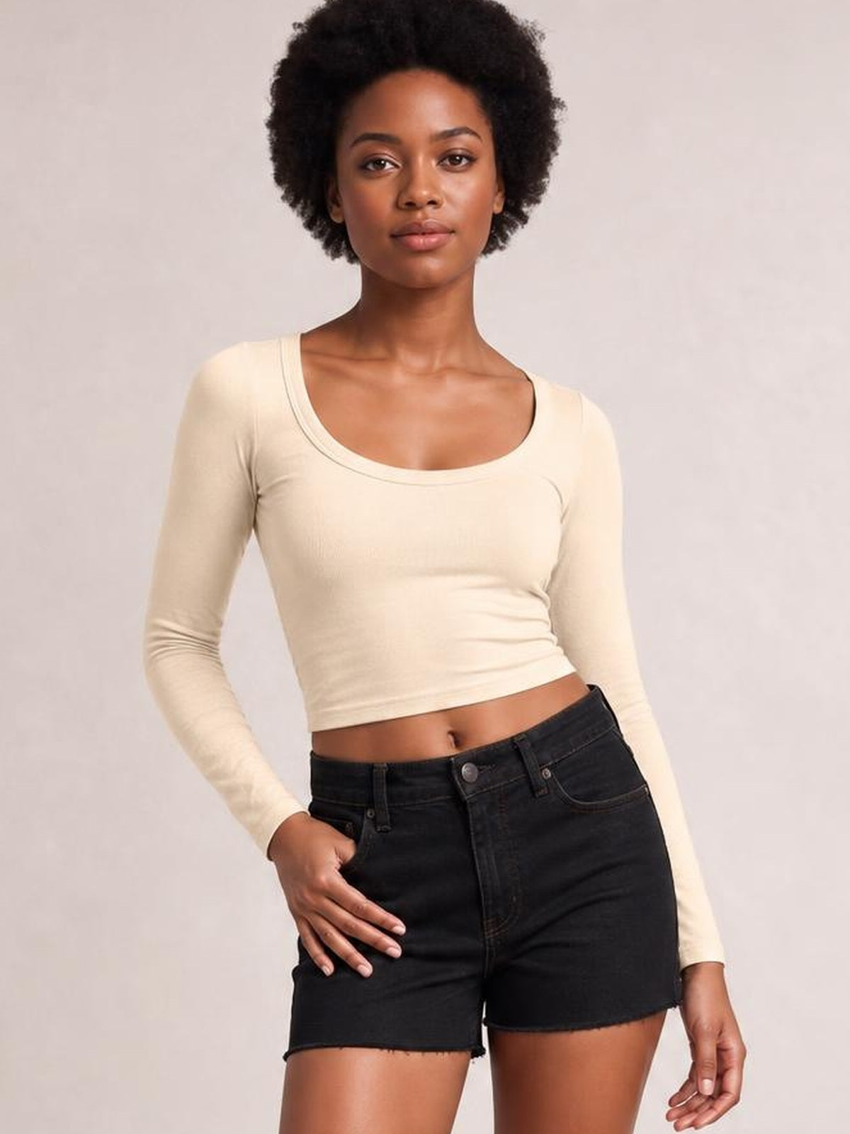 Model wearing a cream scoop neck long sleeve cropped T-shirt with a fitted silhouette and gently curved neckline, posed with one hand on her hip.