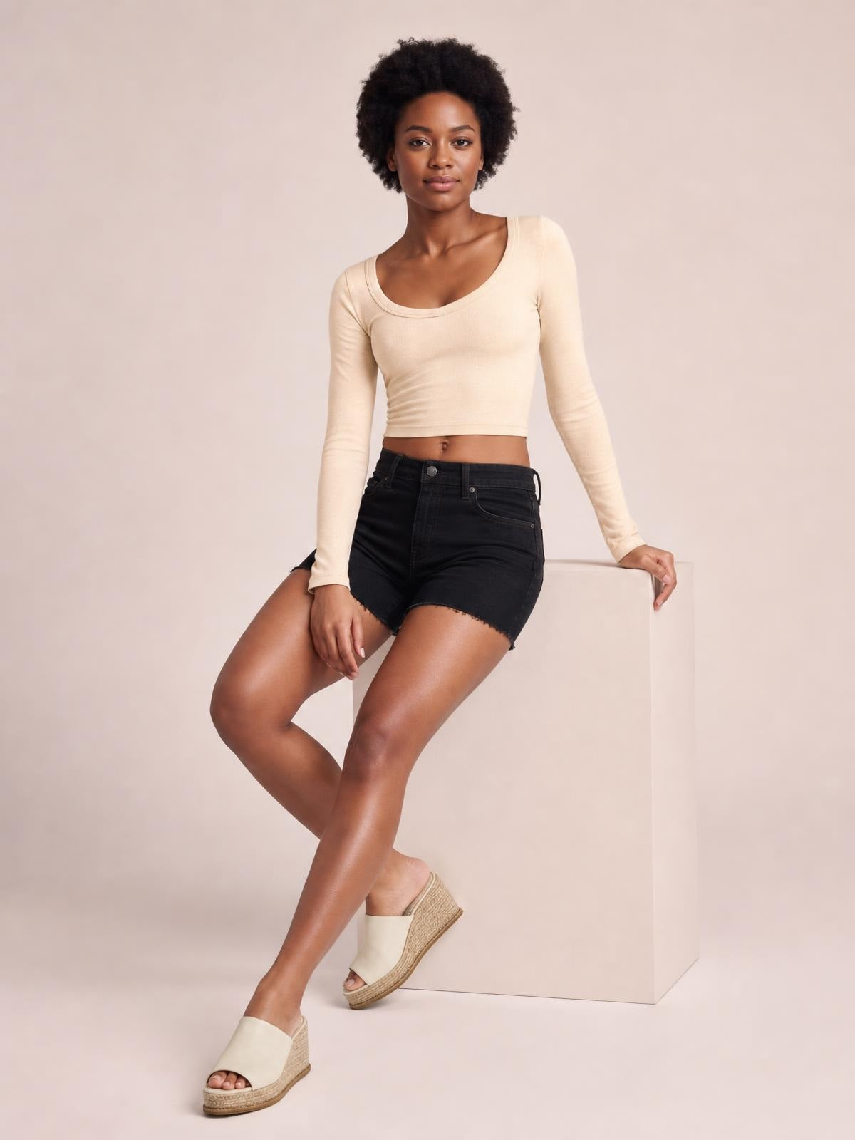 Model wears an ivory scoop neck long sleeve cropped T-shirt with a fitted silhouette and clean bound neckline, seated with legs crossed.