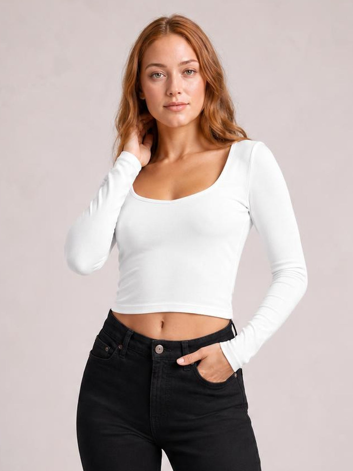A model wears a white scoop-neck long-sleeve cropped T-shirt with a fitted silhouette and smooth hem, touching her hair with one hand and resting the other in her pocket.
