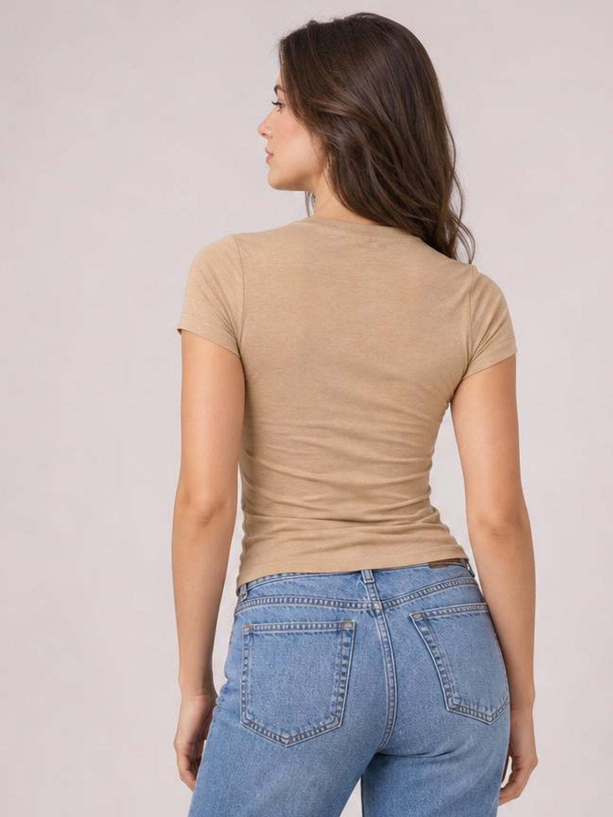 A model stands facing away in a light beige scoop neck short sleeve T-shirt with a close-fitting silhouette and subtle waist ruching.