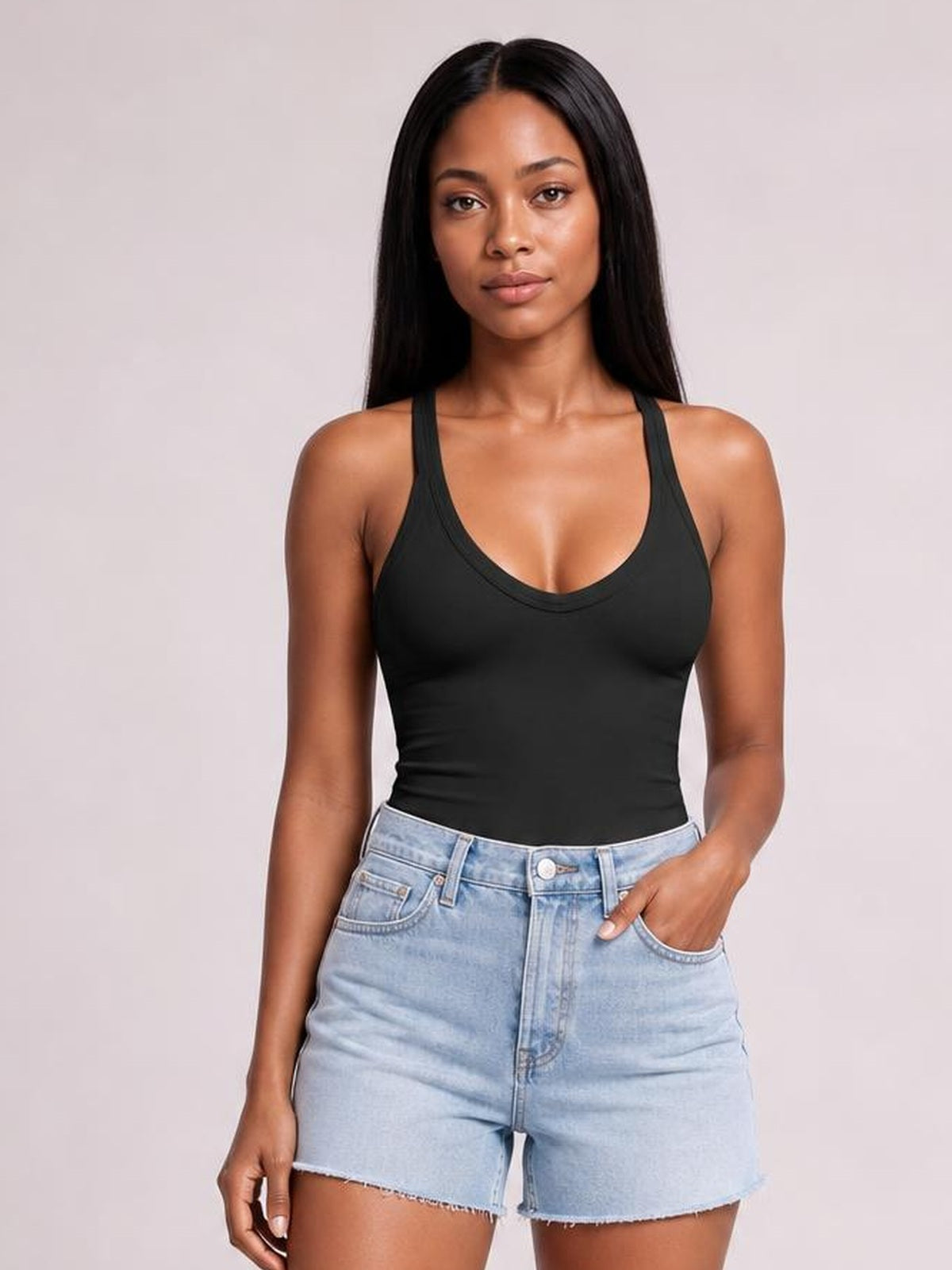 Model wears a black scoop neck wide strap tank with a fitted silhouette and smooth opaque construction, posing with one hand in her denim shorts pocket.