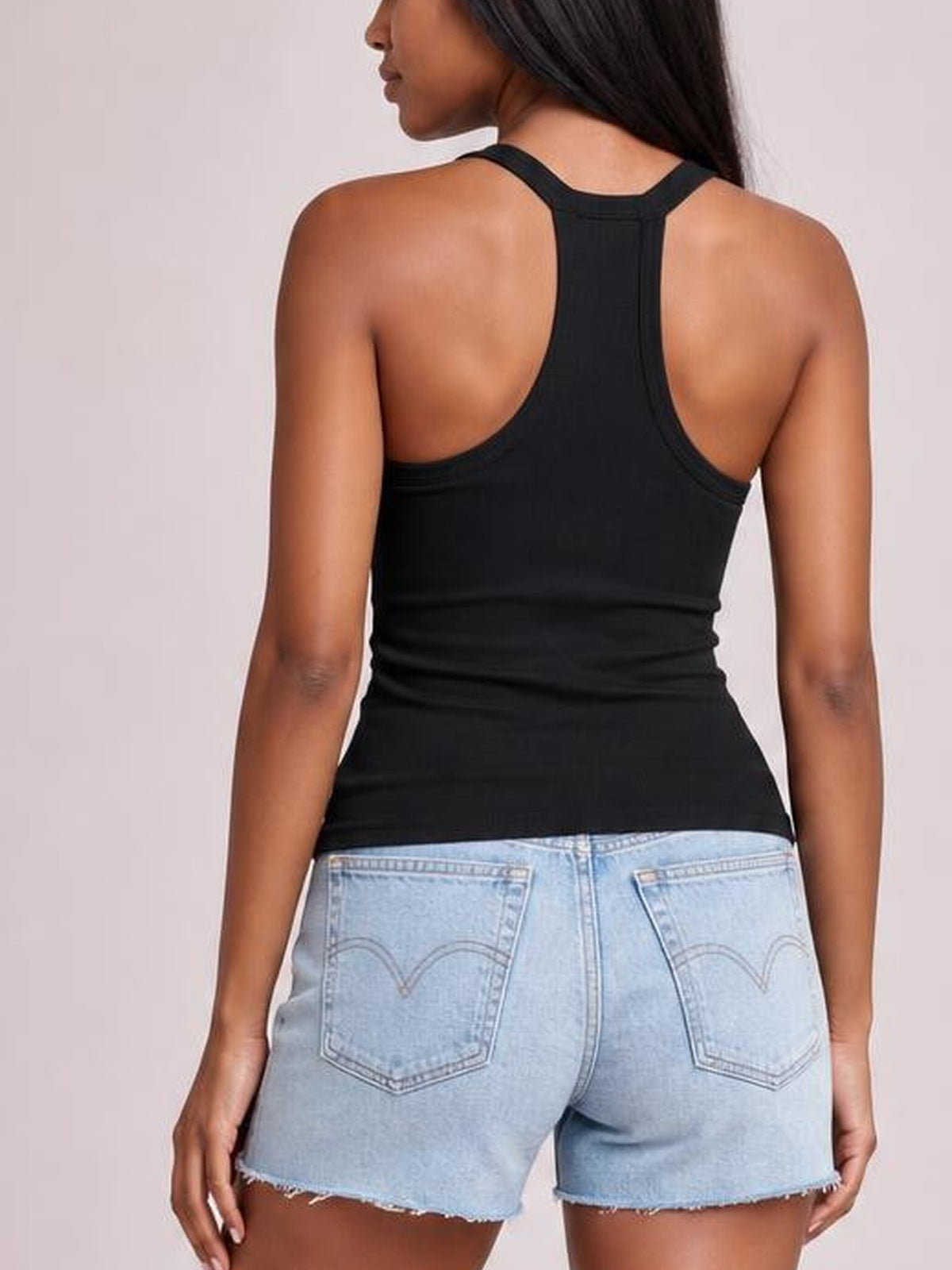 A model wears a fitted black scoop neck tank with wide straps and a racerback-style back, shown from behind with arms resting at her sides.