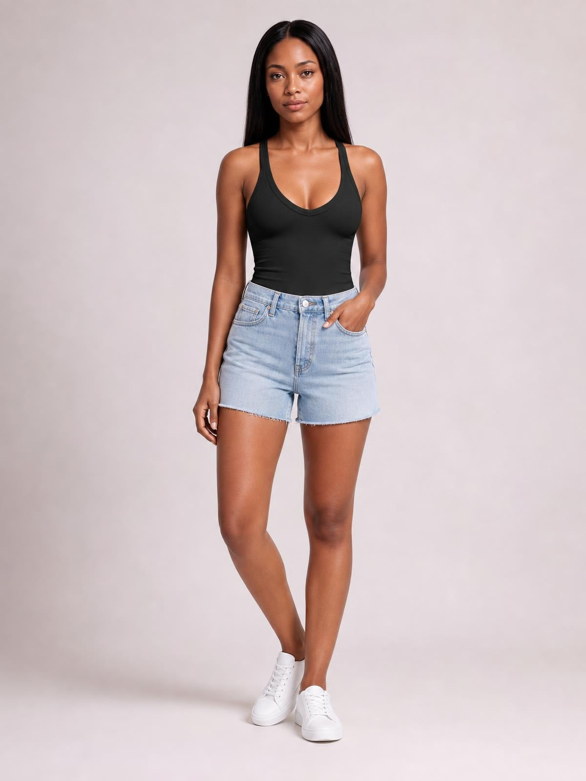Model poses in a black scoop neck tank with wide straps and a fitted silhouette, paired with light-wash denim shorts and white sneakers.