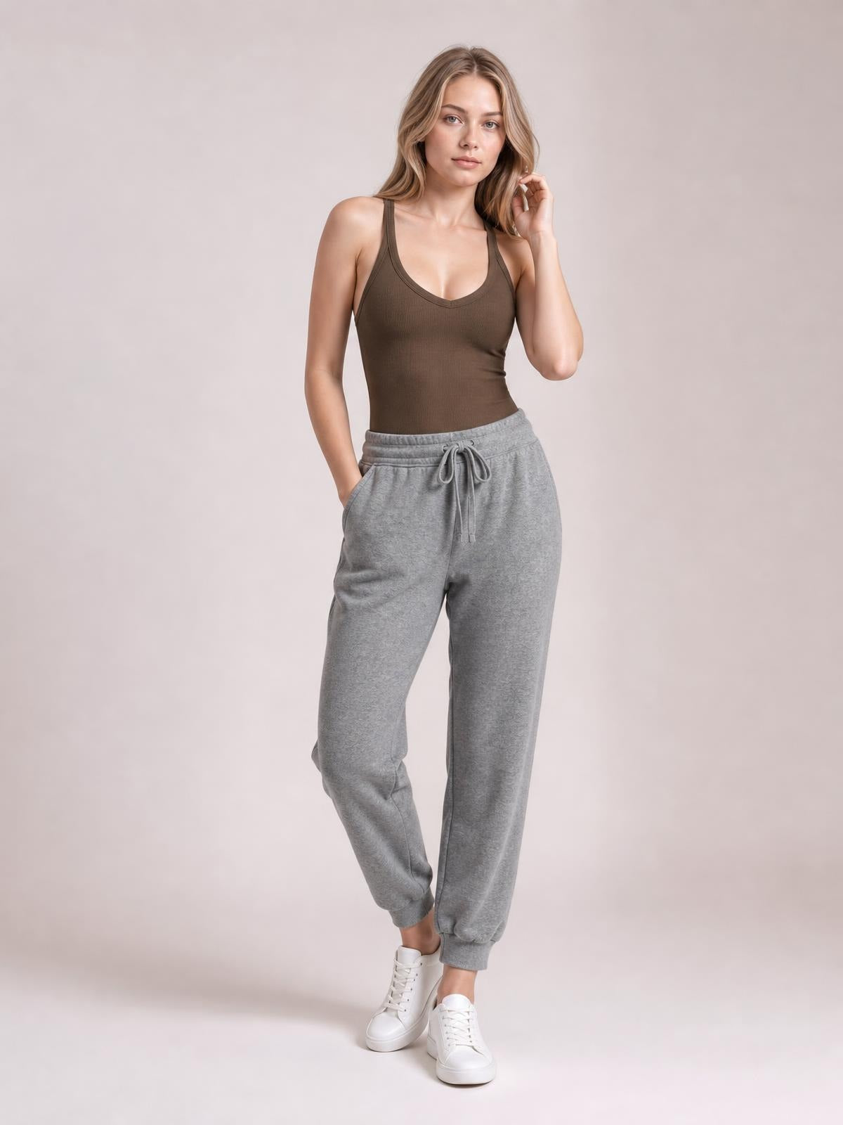 Model wearing a brown scoop neck wide strap tank with a fitted, ribbed silhouette, posing with one hand in a pocket and the other touching her hair.