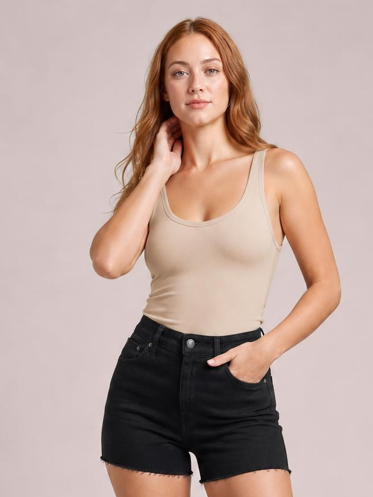 Model poses with one hand at her neck while wearing a beige scoop neck tank with wide straps, a fitted silhouette, and double-stitched neckline.
