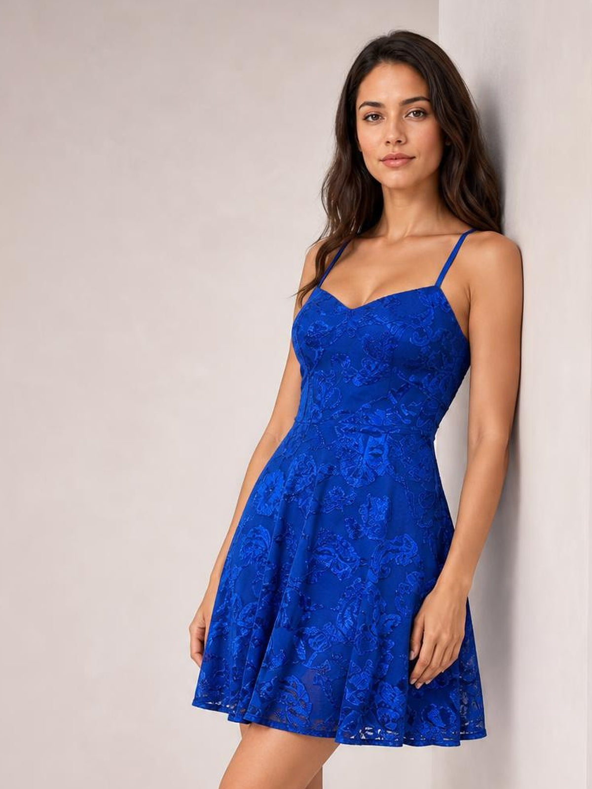 A model leans against a wall wearing a royal blue sequin-embellished mini dress with thin straps, a fitted mid-waist bodice, and a flared skirt.