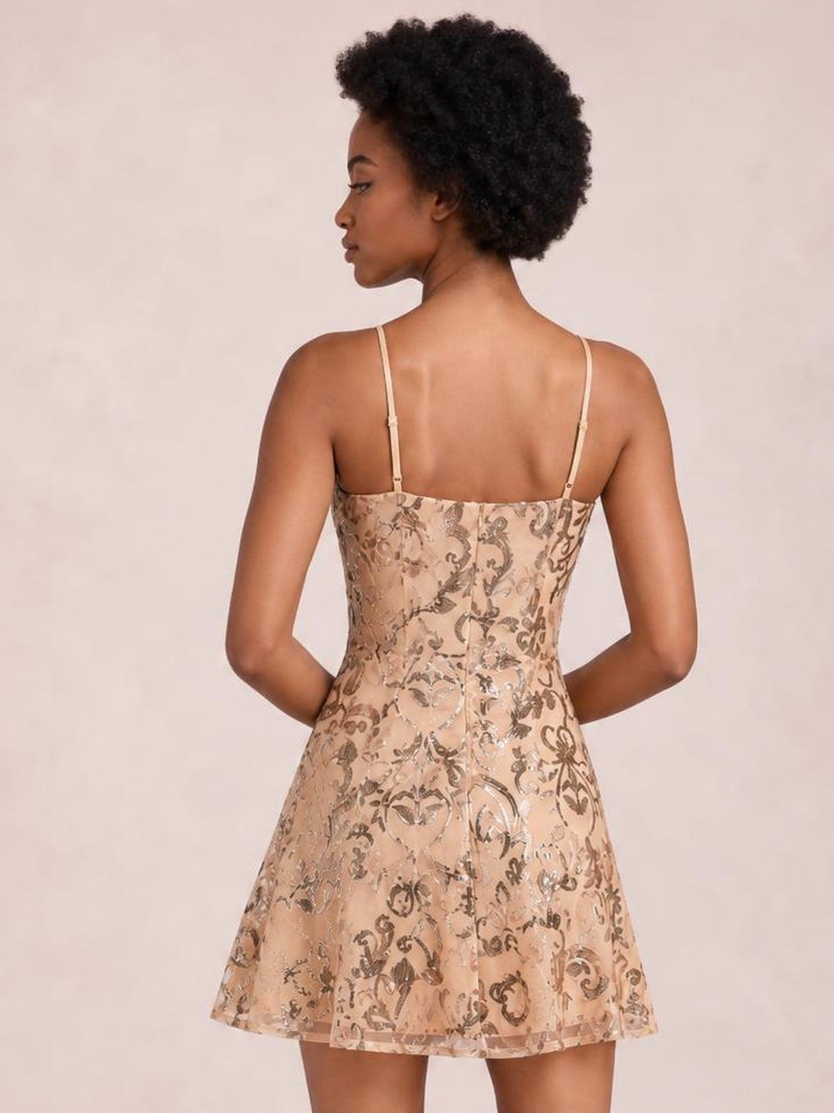 Model poses with hands on hips, showing the back of a champagne sequin embellished mini dress with thin straps, fitted bodice, and flared skirt.