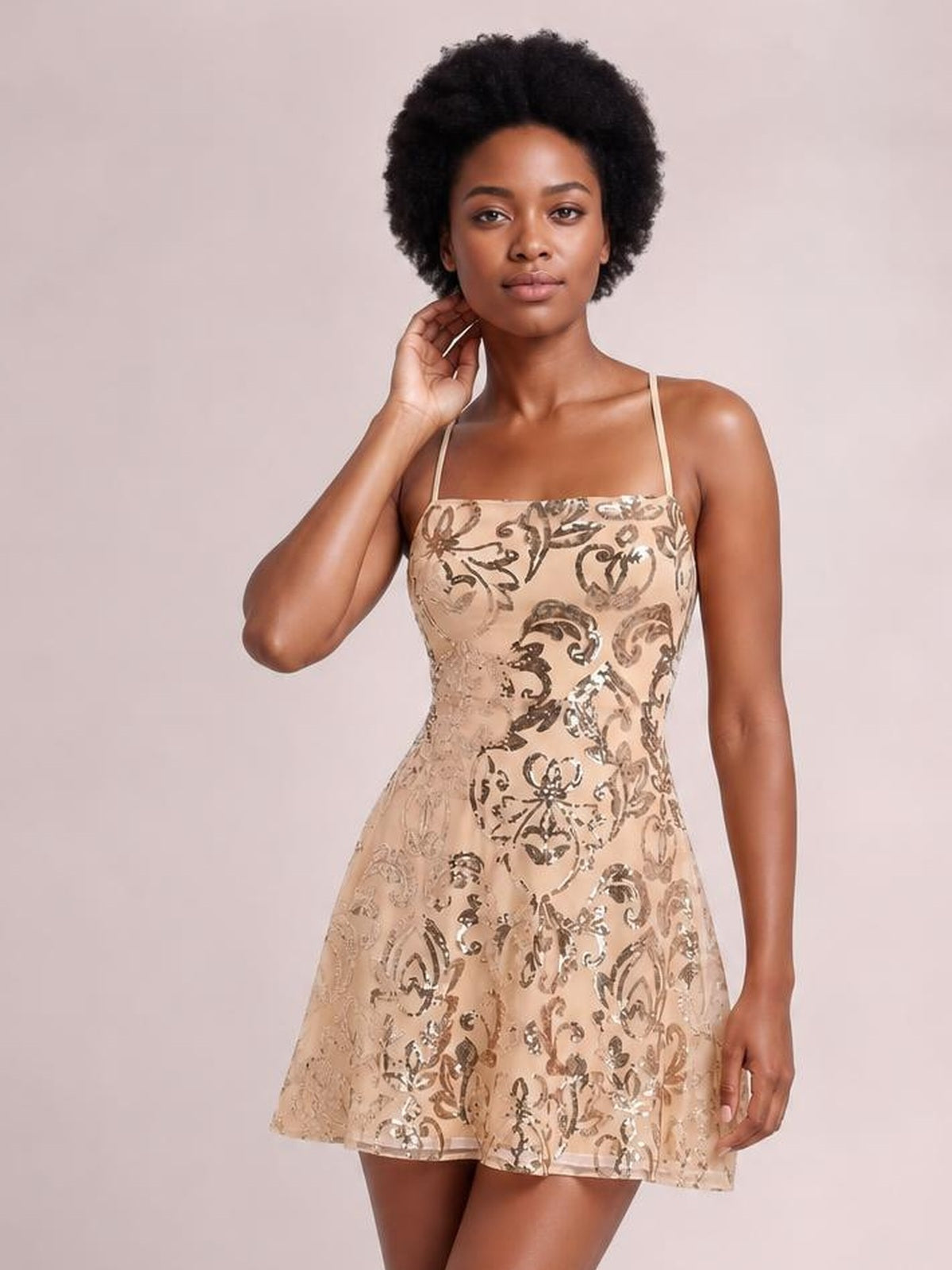 A model poses in a champagne sequin embellished mini dress with thin straps, a square neckline, fitted mid-waist bodice, and flared skirt.
