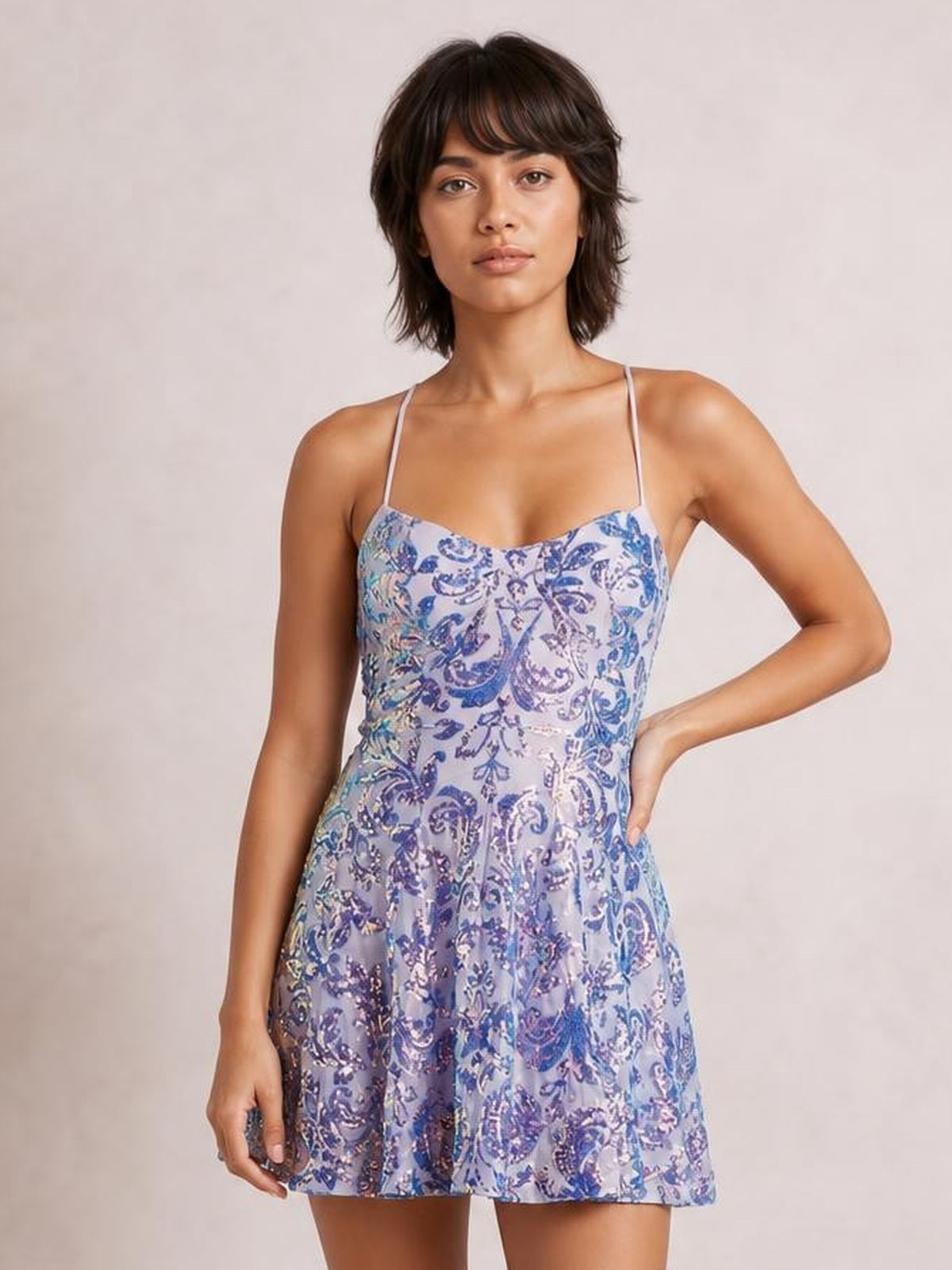 Model poses with one hand on her hip in a lavender-blue sequin embellished mini dress featuring thin straps, a scoop neckline, fitted bodice, and flared skirt.