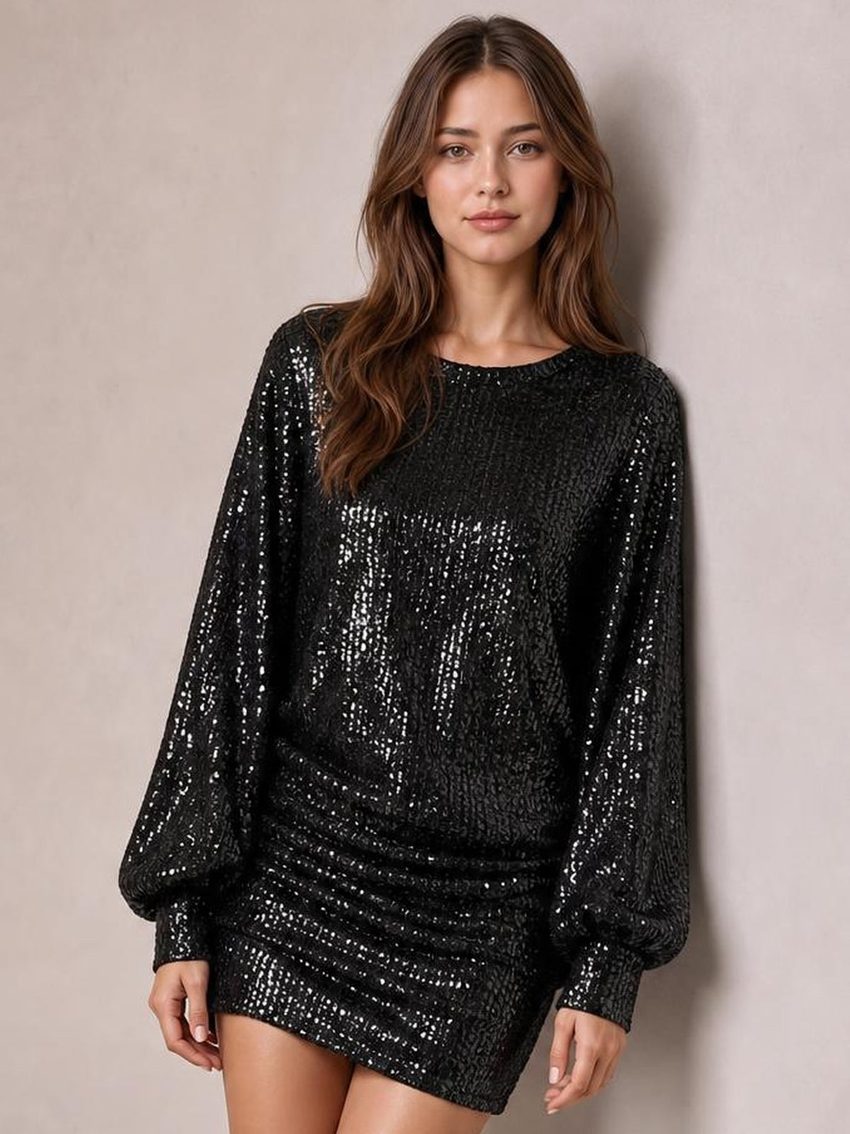 Model poses standing in a black sequin mini dress with long sleeves, a round neckline, relaxed silhouette, and ruched detailing at the waist and hips.