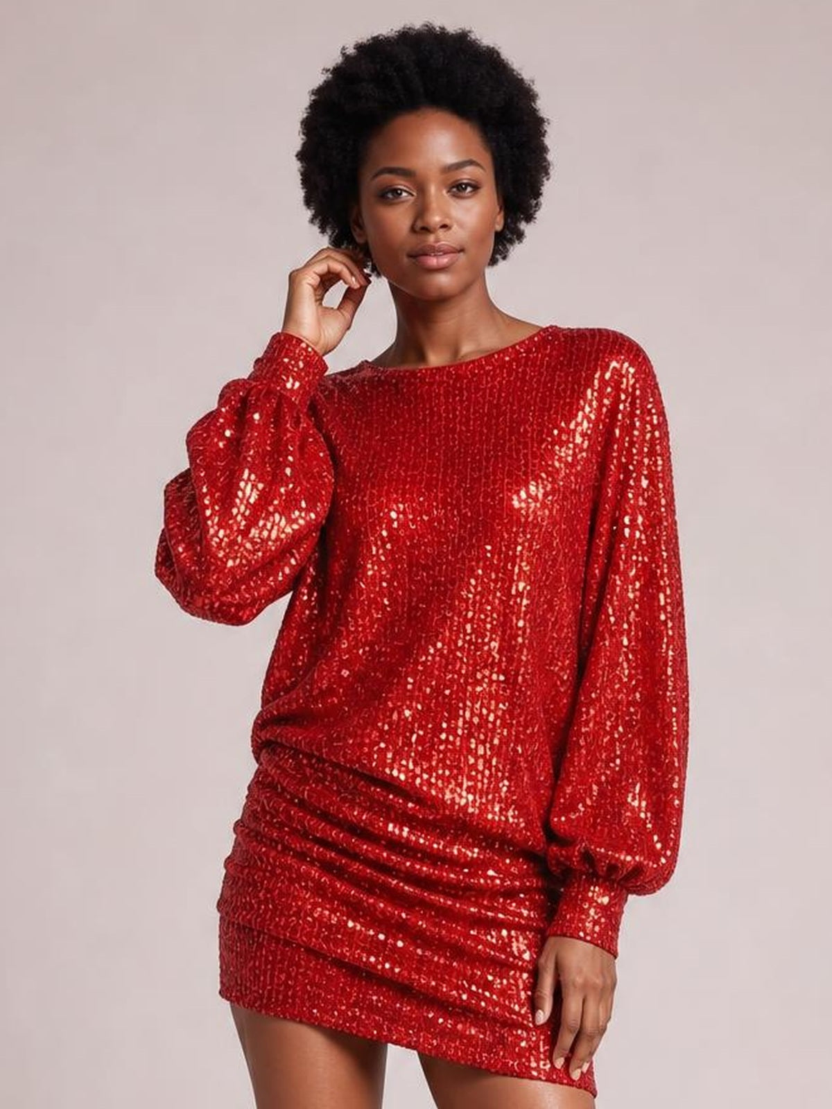 Model poses with one hand near her face in a red sequin long sleeve mini dress featuring a relaxed fit, round neckline, and ruched skirt.